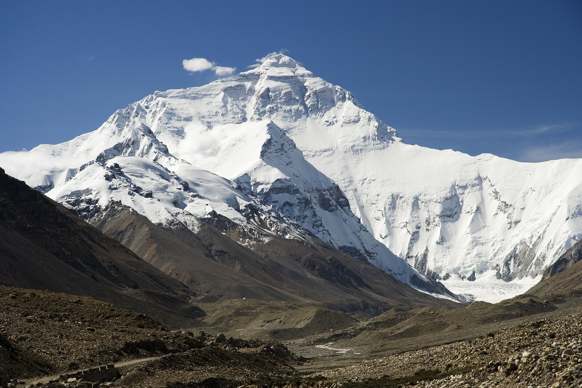 Zdjęcie przedstawia pasmo górskie. To fragment najwyższych gór świata Himalajów. Pasmo górskie pokryte jest śniegiem. W środkowej części pasma najwyższy szczyt w kształcie trójkąta. To Mount Everest, najwyższa góra świata. Skalisty, pokryty śniegiem i oświetlony promieniami słonecznymi szczyt góry kontrastuje z kamienistym, brązowym terenem rozpościerającym się u stóp Mount Everestu. W dole ilustracji widoczne ścieżki. Niebo błękitne.