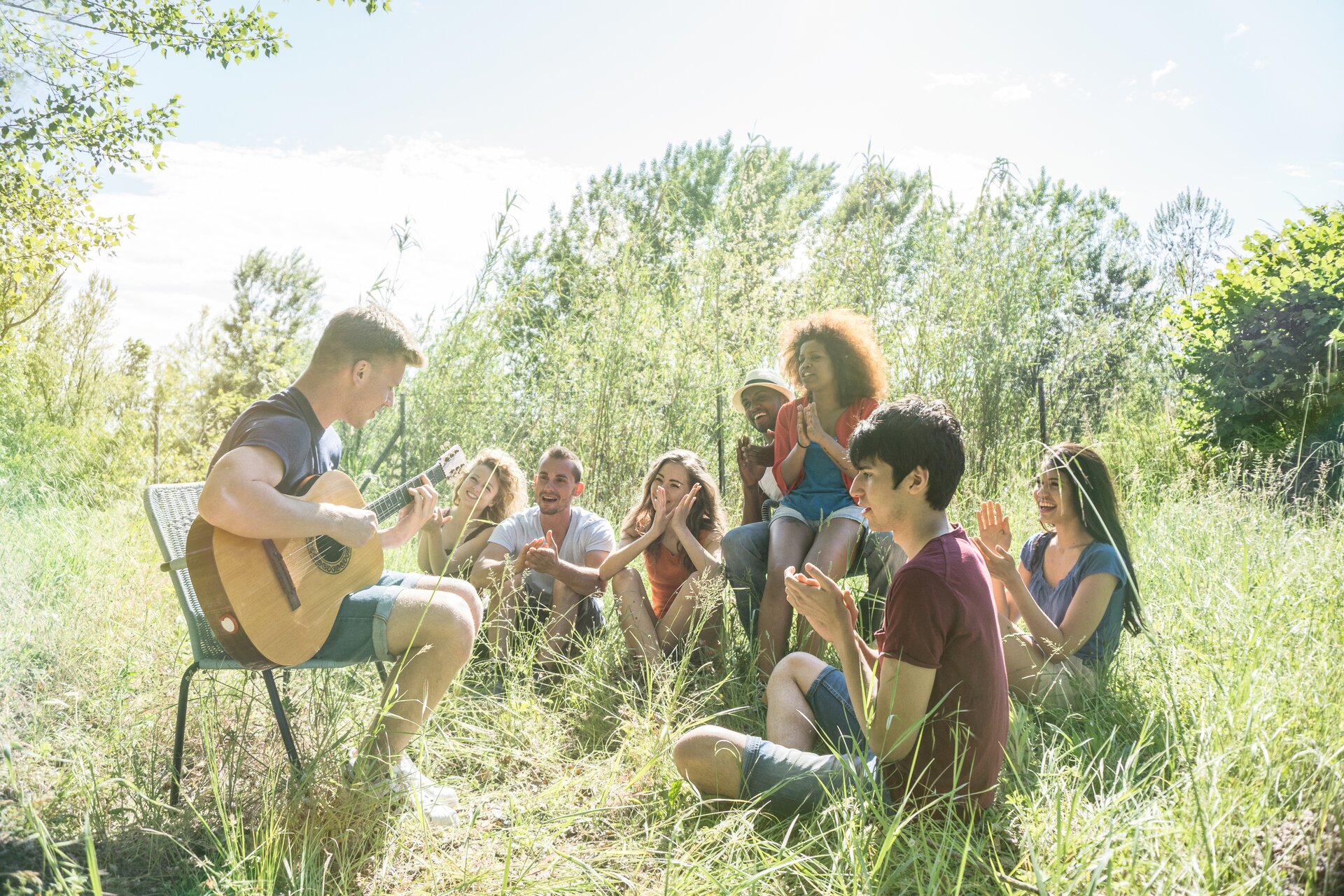 Ilustracja przedstawia grupkę przyjaciół na łonie natury. Jeden z mężczyzn po lewej stronie siedzi na krześle ogrodowym i gra na gitarze, wokół niego znajdują się nastolatkowie, którzy się uśmiechają i klaszczą.