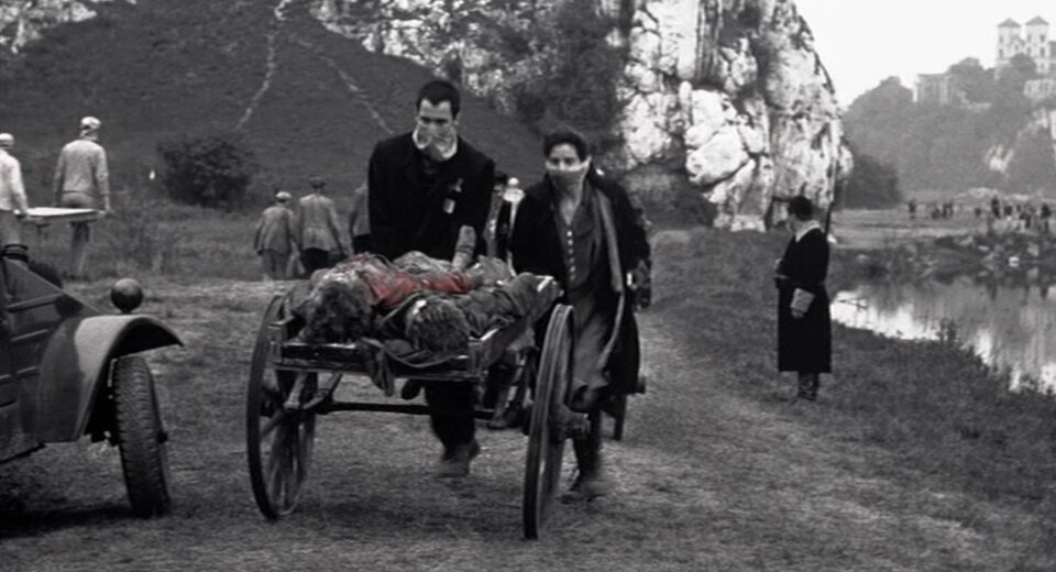 Zdęcie czarno‑białe przedstawia bulwar nad rzeką. Idzie nim, pchając przed sobą wózek, kobieta i mężczyzna. Na wózku znajdują się ciała ludzi. Jednym z nich jest ciało dziewczynki w czerwonym płaszczu. 