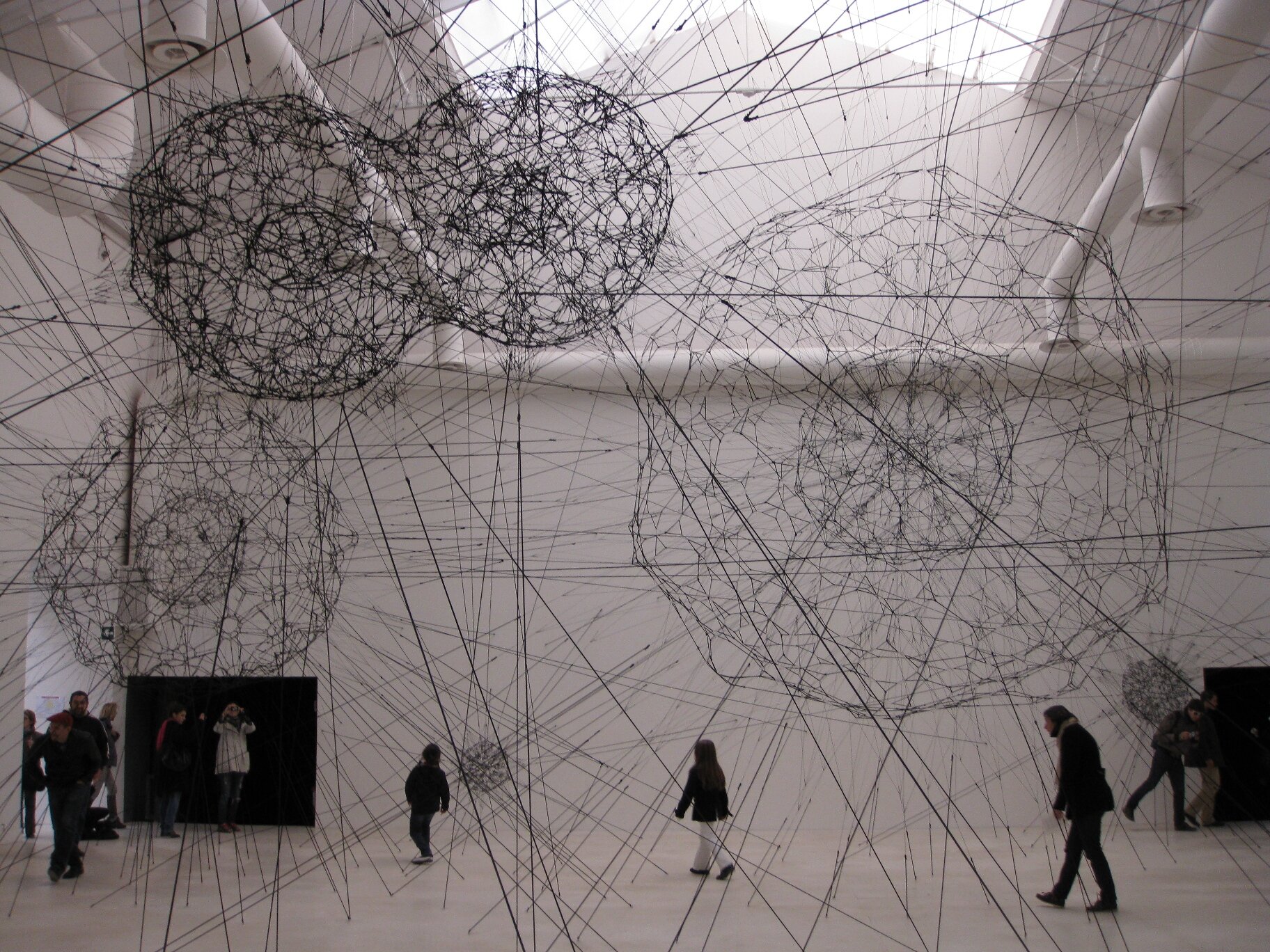 Zdjęcie ukazuje wnętrze wysokiego pomieszczenia, w którym znajduje się kilka osób w ciemnych ubraniach. Postacie są w jasnym, wysokim pomieszczeniu o jasnej podłodze. Nad ludźmi znajdują się 4 duże, ażurowe kule w kolorze czarnym. Niżej są 2 o mniejszej średnicy. Dodatkowo w przestrzeni pomieszczenia umieszczona została konstrukcja zbudowana z różnego rodzaju lin przybierająca kształt sieci. Liny biegną wzdłuż i wszerz pomieszczenia, kotwy mocujące konstrukcję widoczne są także na suficie i posadzce.