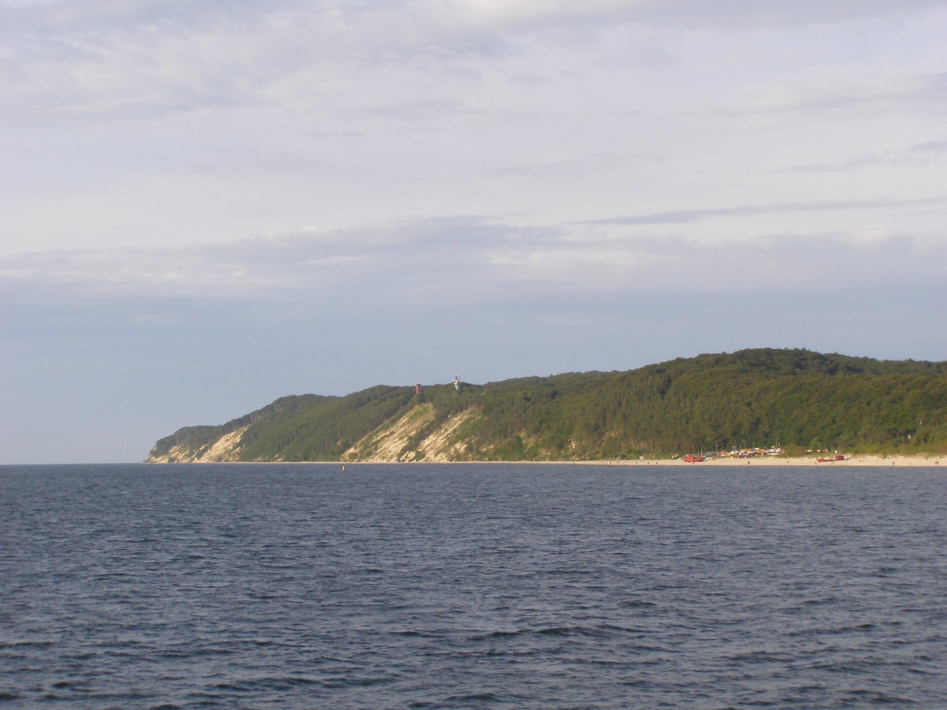 Fotografia, na której na pierwszym planie jest rozległe morze, a dalej rozległy i masywny klif. Jego zbocza są dość strome, w większości porośnięte drzewami. Gdzieniegdzie odsłonięte zbocza w kolorze piaskowym, u dołu piaszczysta plaża, po której spacerują ludzie.