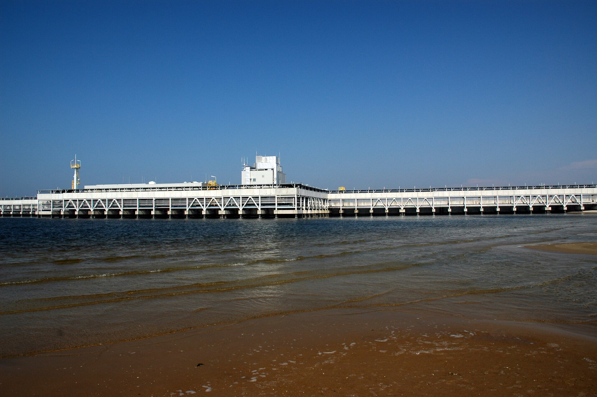 Na zdjęciu widać z oddali podłużną budowlę, sięgającą daleko w głąb morza. Konstrukcja jest jednolita, z odległej perspektywy mogłaby się kojarzyć z molo. Na pierwszym planie widzimy morską plażę. Jasna płytka woda przy brzegu, ciemniejsza i głębsza w miarę posuwania się w kierunku budowli.