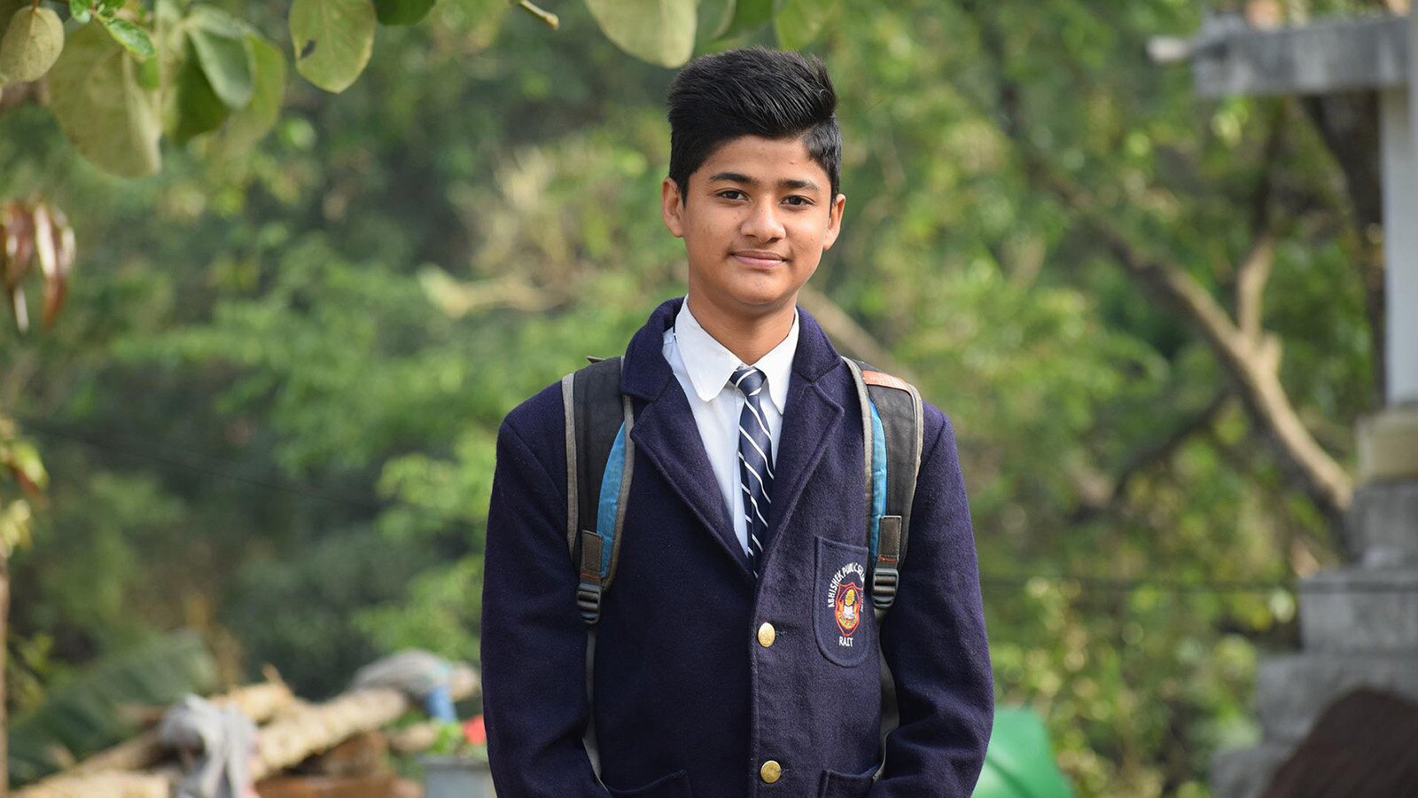 Zdjęcie przedstawia nastolatka ubranego w marynarkę z haftowanym wzorem na kieszeni oraz białą koszulę i krawat. 