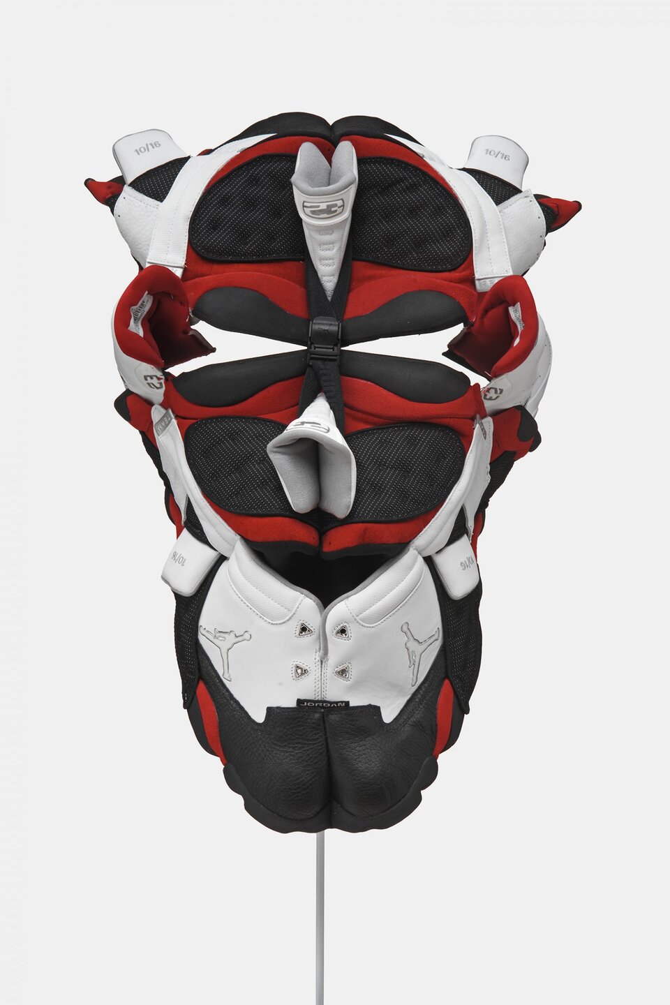 Dzieło widoczne na fotografii wykonano z elementów obuwia sportowego. Kształtem przypomina maskę z otworami na oczy oraz elementem upodobnionym do nosa. Pod nim, na dole dzieła zauważalne jest logo modelu obuwia - mężczyznę wyskakującego z piłką koszykową. Logo oraz części dzieła są białe, jednak widoczne są również czerwone oraz czarne elementy. U góry, po obu stronach oraz u dołu, można zauważyć liczby - 10/16. Dodatkowo, u góry pracy widnieje liczba 23. Dzieło jest symetryczne. Z prawej oraz z lewej strony wyróżniają się elementy z metką, które zdają się być zapiętkiem buta.
