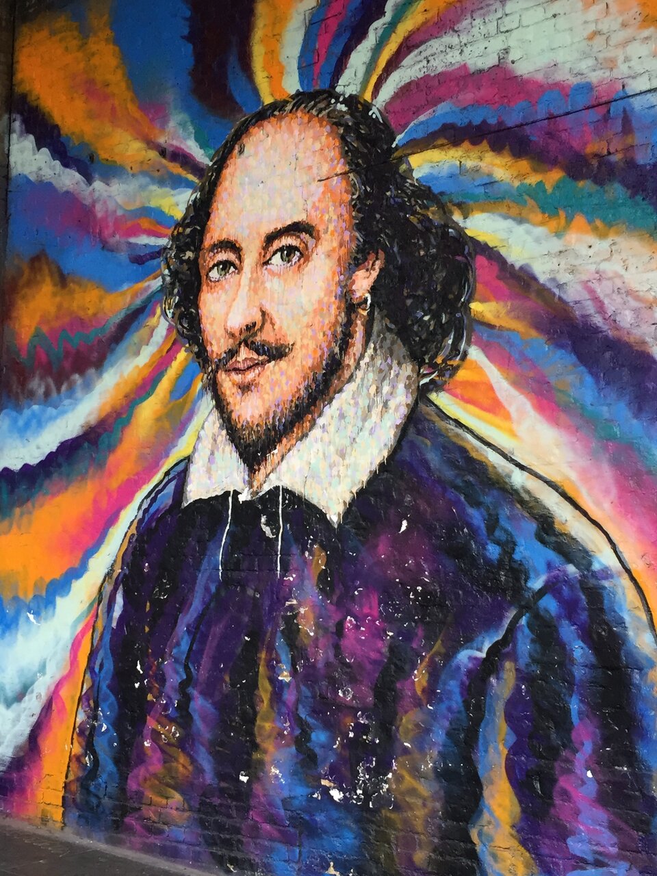 Zdjęcie przedstawia mural z portretem mężczyzny. Mężczyzna jest młody. Ubrany jest zgodnie z siedemnastowieczną modą angielską. W kaftan i koszulę z szerokim kołnierzem. Mężczyzna ma długie, układające się, ciemne włosy. Ma ciemne brwi i oczy. Jego twarz zdobi niewielki wąs i krótka broda. Twarz mężczyzny jest pociągła. Od głowy mężczyzny rozchodzą się promieniście różnokolorowe pasy.