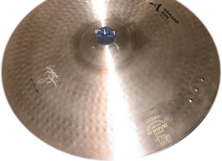 Zdjęcie przedstawia okrągły talerz perkusyjny w kolorze jasnego złota, z lekko wypukłą, okrągłą częścią pośrodku, w której umieszczone jest czarne pokrętło motylkowe. W górnej i dolnej części talerza czarne napisy i logo.