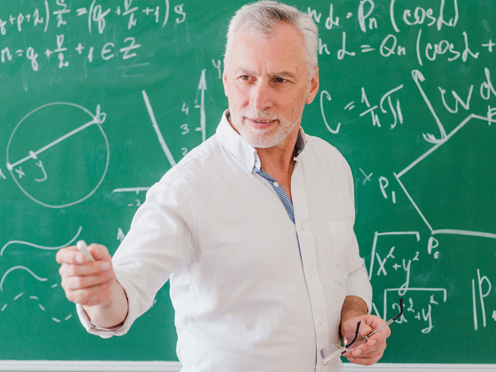 Zdjęcie przedstawia starszego mężczyznę, w ujęciu od pasa w górę, który stoi przed zieloną tablicą. Mężczyzna ma krótkie, siwe włosy i jest ubrany w białą koszulę. W lewej ręce przyciśniętej  do brzucha trzyma okulary korekcyjne, zaś w prawej ręce wyciągniętej do przodu trzyma kredę. Na tablicy widoczne są różne wzory matematyczne zapisane białą kredą.