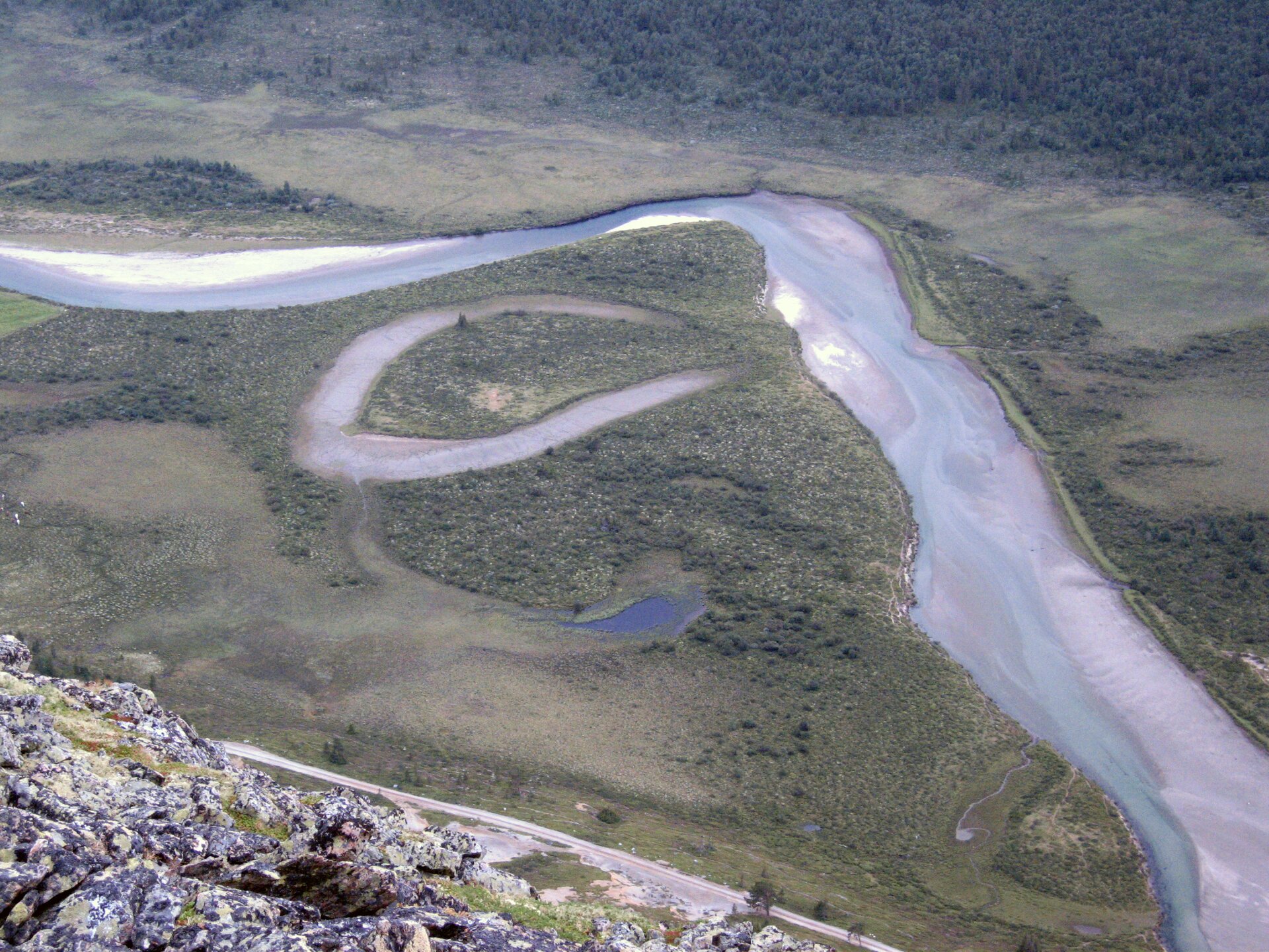 Fotografia prezentuje dolny bieg rzeki – koryto rzeczne jest szerokie, brzegi piaszczyste. Na dnie widać dużo piasku, który sprawia, że rzeka jest płytsza. Obok rzeki widać odcięte od niej zakole w kształcie litery U. Zakole z wodą jest odgrodzone od rzeki lądem.