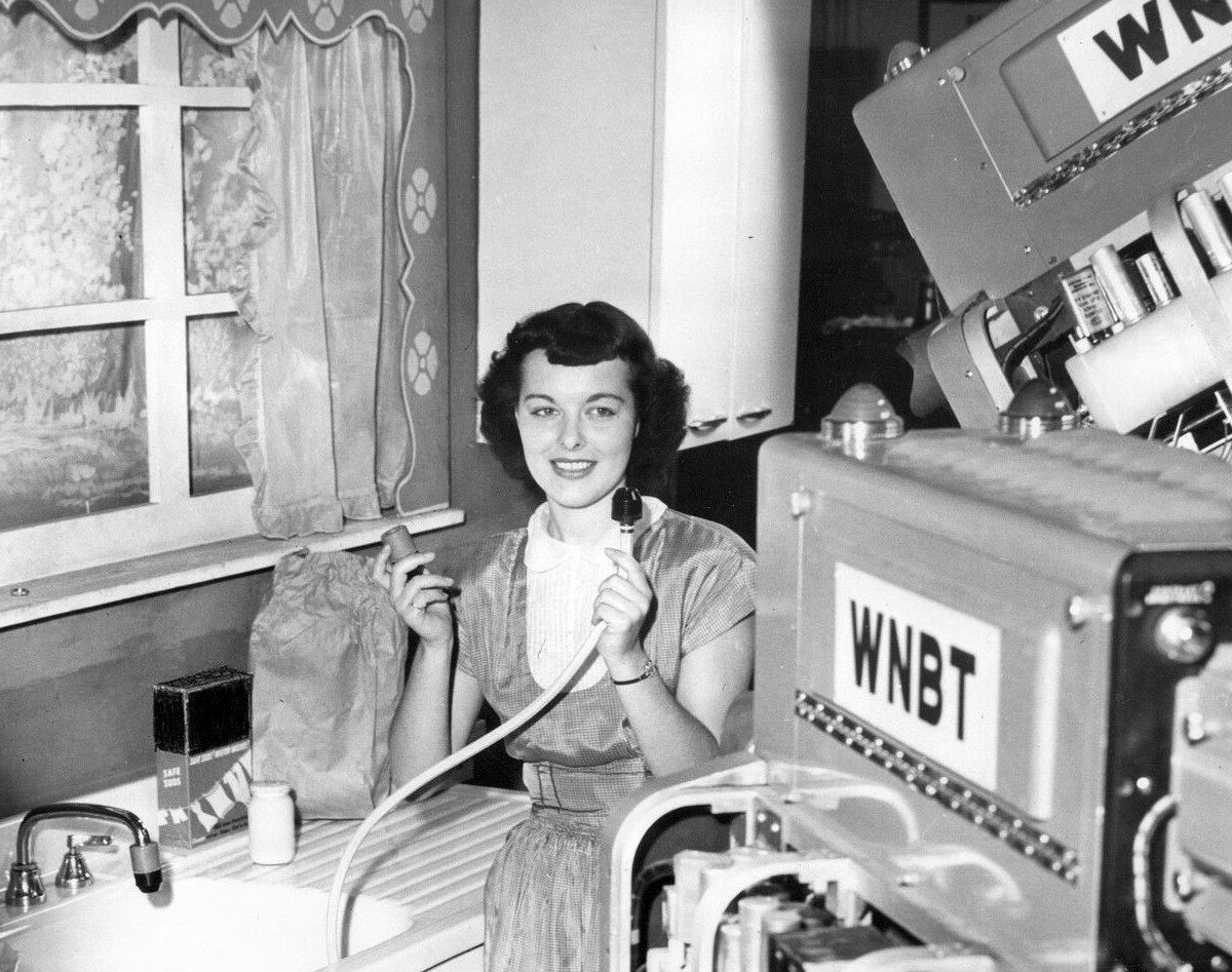 Czarno‑białe zdjęcie przedstawiające stojącą przy zlewozmywaku kobietę, trzymającą w dłoni perlator nakładany na końcówkę kranu napowietrzający strumień wody.