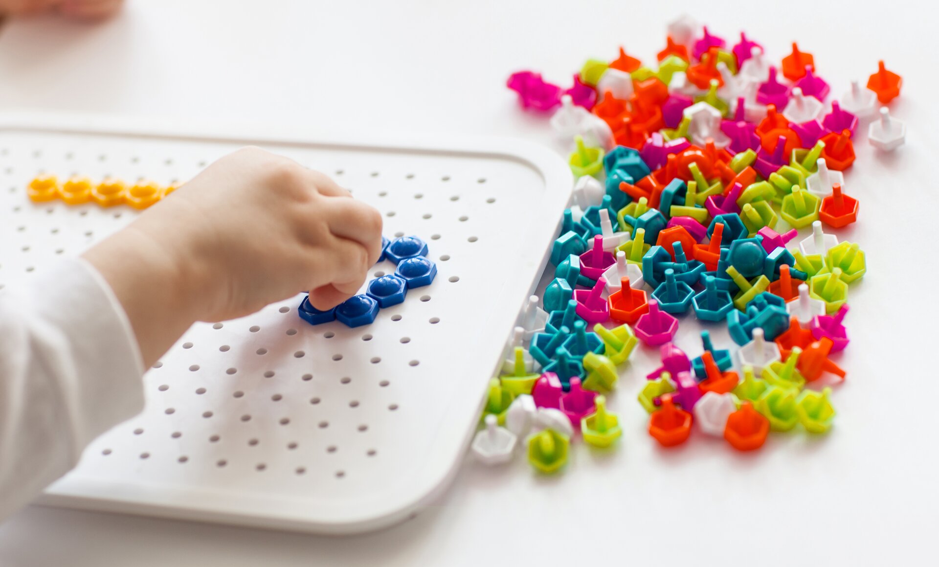 Zdjęcie przedstawia mozaikę wtyczkową, którą układa dziecko. Mozaika to plastikowa prostokątna deska z licznymi dziurkami. Można w nie wkładać małe plastikowe wtyczki, które również widać na zdjęciu. Są to płaskie sześciokąty foremne na krótkim trzonku. Mają różne kolory.