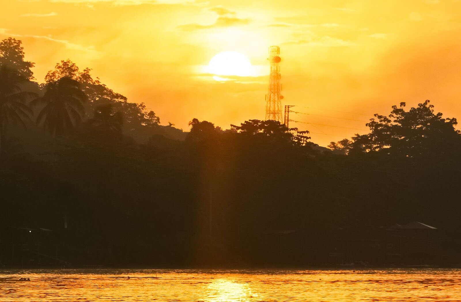 Zdjęcie przedstawia rozpaloną, białą tarczę słońca widoczną na żółto‑pomarańczowym niebie. Poniżej widoczne są ciemne kontury drzew oraz tafla wody, w której odbija się słońce i niebo.