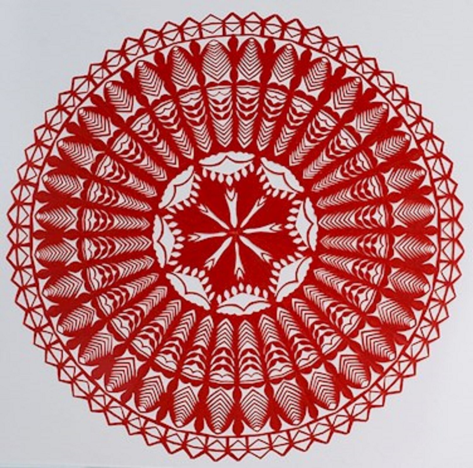 Ilustracja przedstawia kurpiowską gwiazdę. Wzór jest wykonany w kolorze czerwony. Składa się z trzech pierścieni o różnej grubości i kola w środku. Zewnętrzny pierścień dookoła to ułożone obok siebie otwarte koperty. Każdy pierścień ma inny wzór, a środek to gwiazda o ośmiu wierzchołkach.