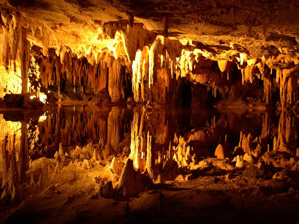 Zdjęcie przedstawia jaskinię krasową. Widoczne są stalaktyty, stalagmity i zbiorniki wodne. Jaskinia jest jasno oświetlona, ma jasnobrązową i złotą barwę. 