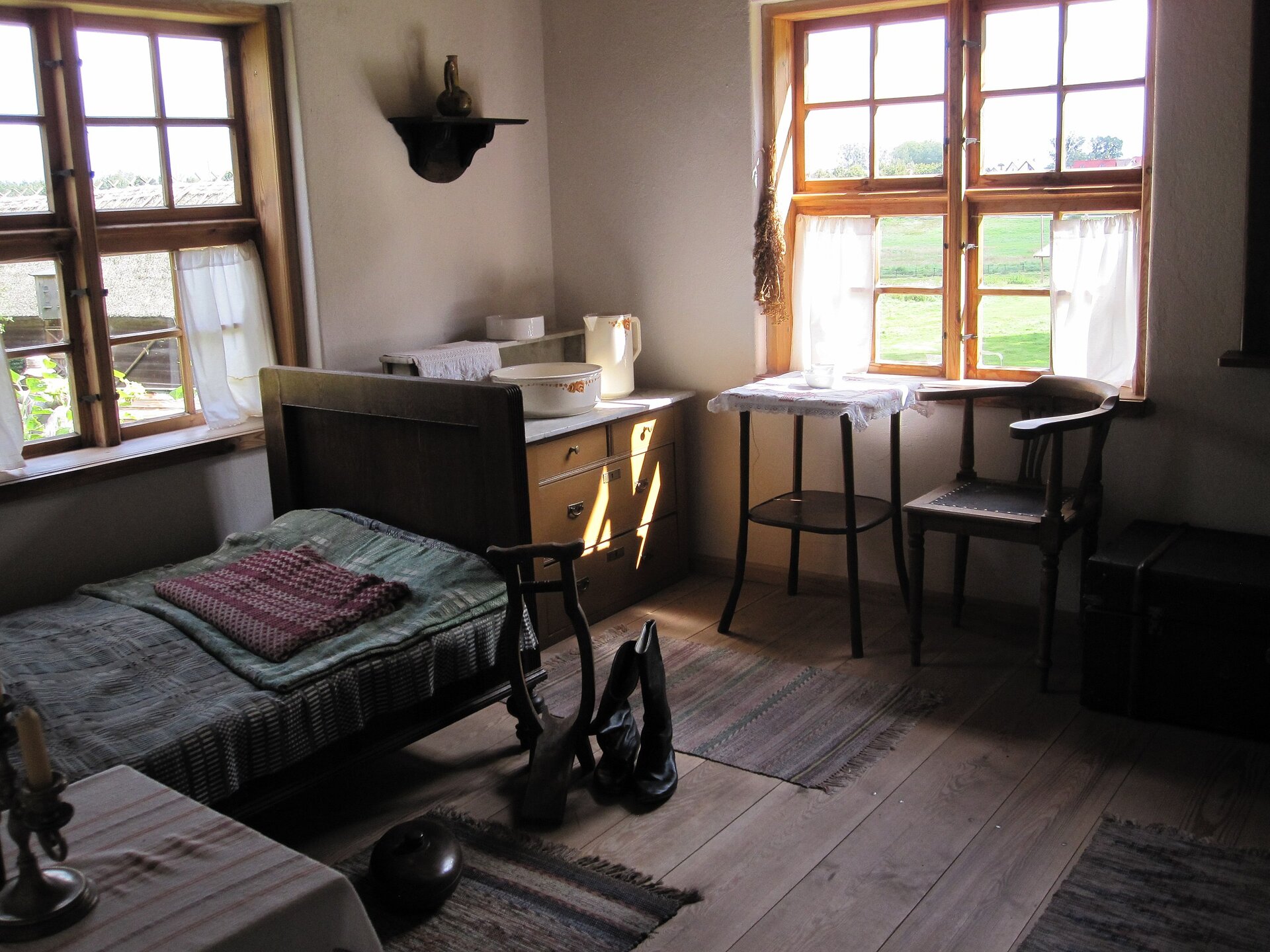 Zdjęcie przedstawia pomieszczenie, w którym po lewej stronie znajduje się niewielki stolik, dalej łóżko, komoda stojąca w rogu, na niej kilka misek. Z prawej strony krzesło i stolik pod oknem. Drugie okno znajduje się nad łóżkiem. 