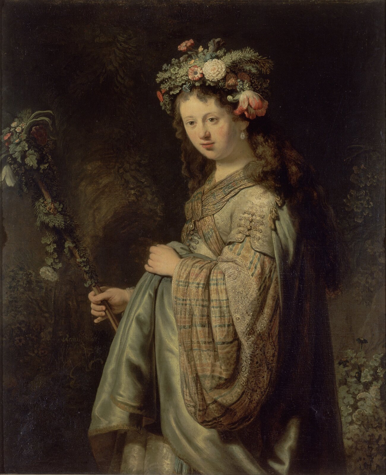 Ilustracja przedstawia obraz Rembrandta van Rijna „Saskia jako Flora”. Na ilustracji znajduje się kobieta w długich, brązowych, kręconych włosach. Na głowie ma wianek z kwiatów. Ubrana jest w szaro-brązowy płaszcz. W dłoni trzyma różdżkę oplecioną kwiatami. Za kobietą znajduje się ciemne tło. 