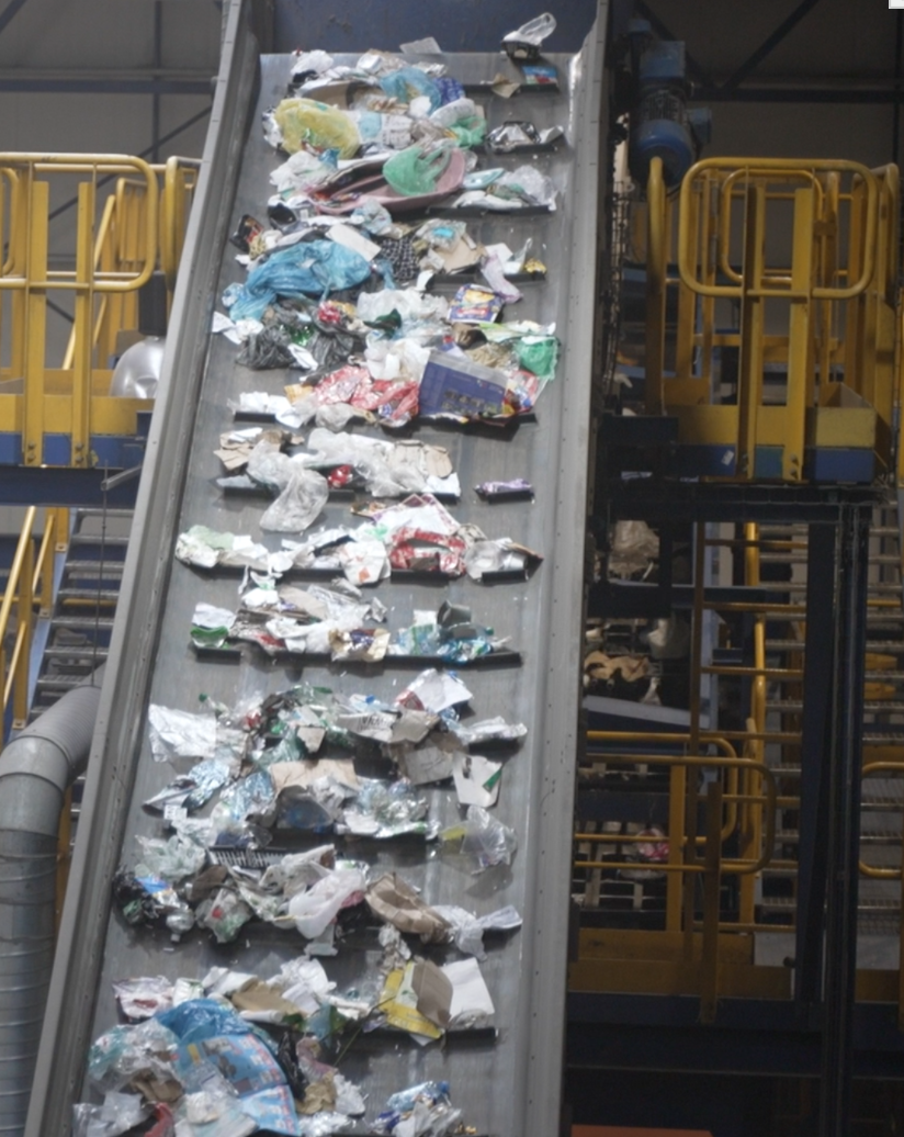 Ilustracja przedstawia transportowanie śmieci, które znajdują się na taśmie w sortowni. 