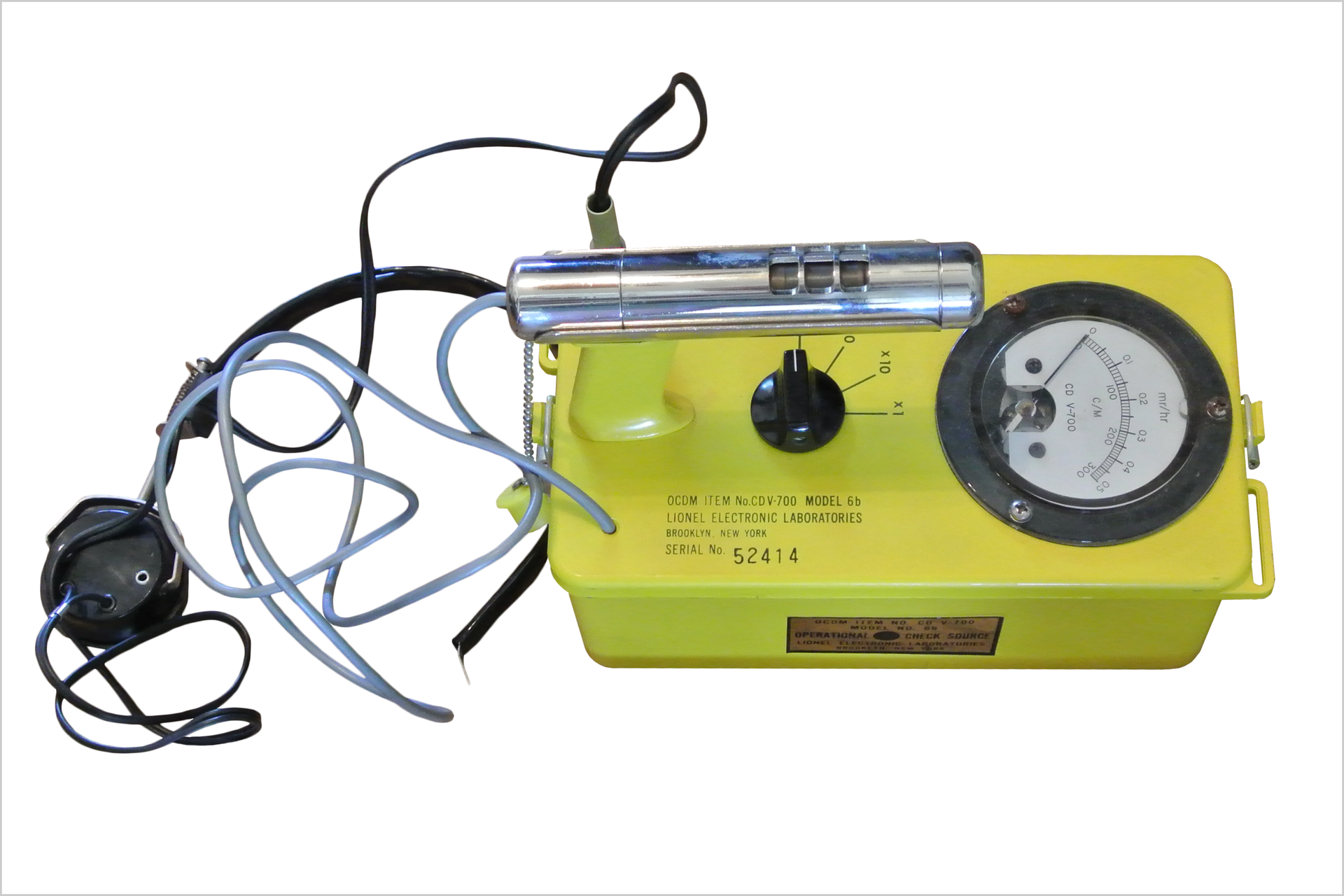 Kolorowe zdjęcie przedstawia licznik Geigera. Zdjęcie wykonane z góry. Po prawej żółte metalowe pudełko. Na górze pudełka znajdują się (od prawej): wskaźnik promieniowania jądrowego ze skalą liczbową, czarne plastikowe pokrętło na środku, po lewej detektor w kształcie rurki. Po lewej stronie zdjęcia kilka kabli i przewodów. Jeden przewód podłączony jest do obudowy drugi do detektora w kształcie rurki.