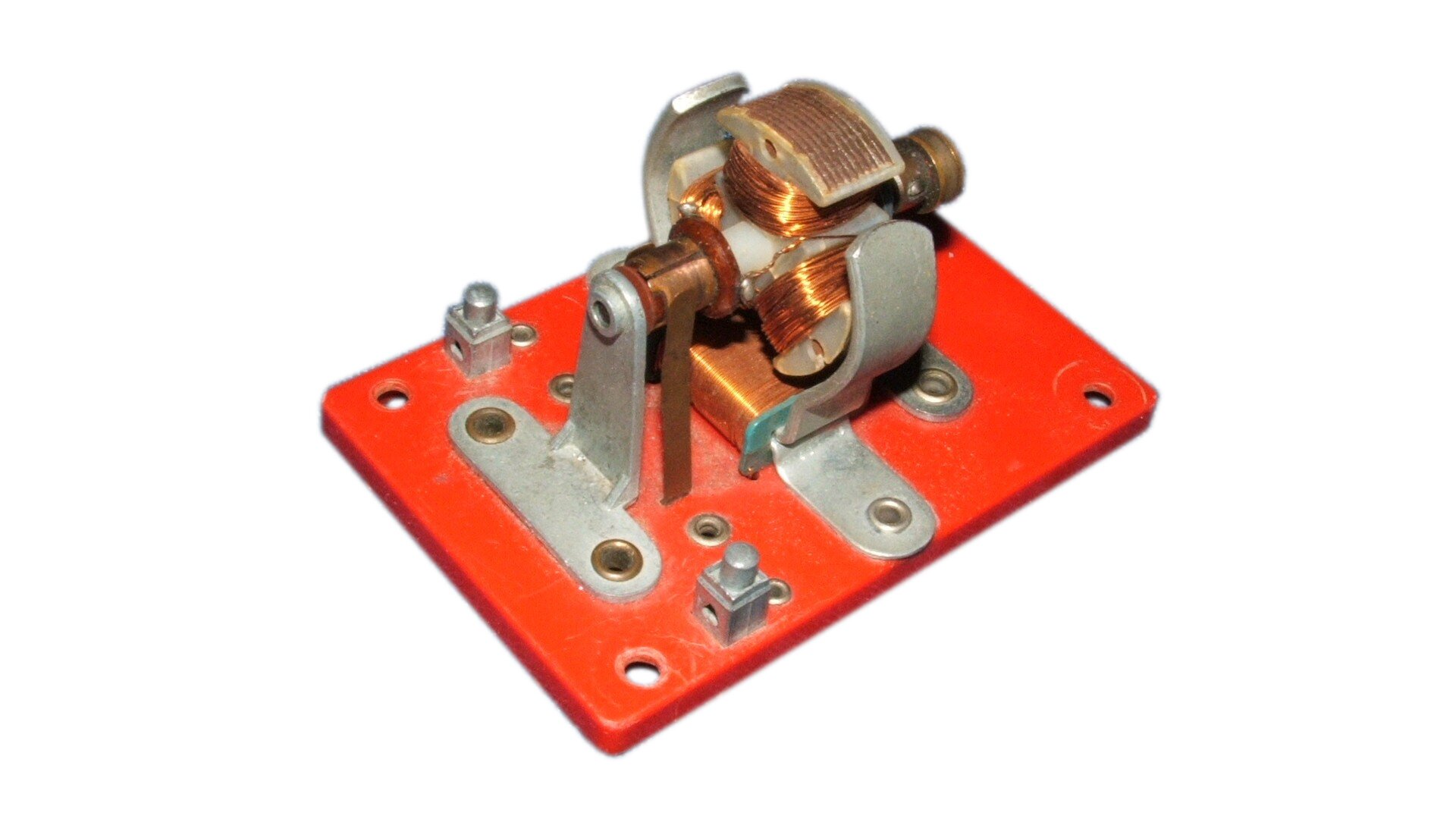 Zdjęcie przedstawia model silnika elektrycznego. Do czerwonej podstawki przykręcono kilka elementów z nawiniętymi drutami miedzianymi. Trzy z tych elementów przytwierdzone są do środka osi tak, że zwoje znajdują się prostopadle do osi. Pod nimi przytwierdzony do płytki prostokątny element, również z nawiniętym drutem. Po obu stronach środka osi, nieznacznie oddalone od zwojów z drutem, łukowato zagięte szare magnesy. 