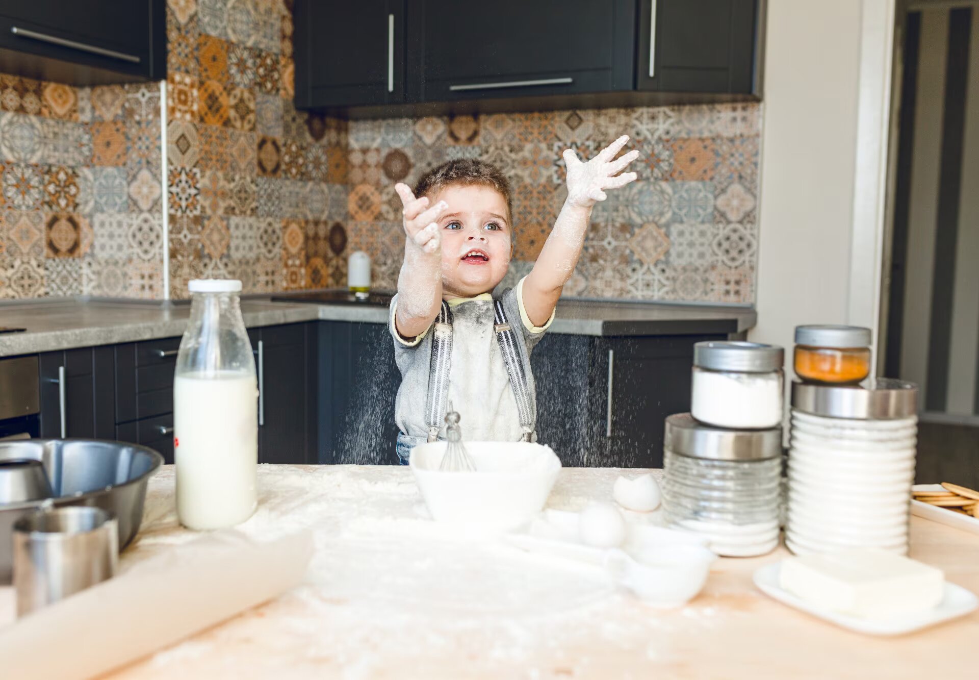 Zdjęcie przedstawia chłopca w wieku przedszkolnym, unoszącego dłonie w górę, który stoi w kuchni przed blatem z mąką, jajkami i różnymi składnikami w szklanych słoiczkach. Chłopiec cały jest pobrudzony mąką.