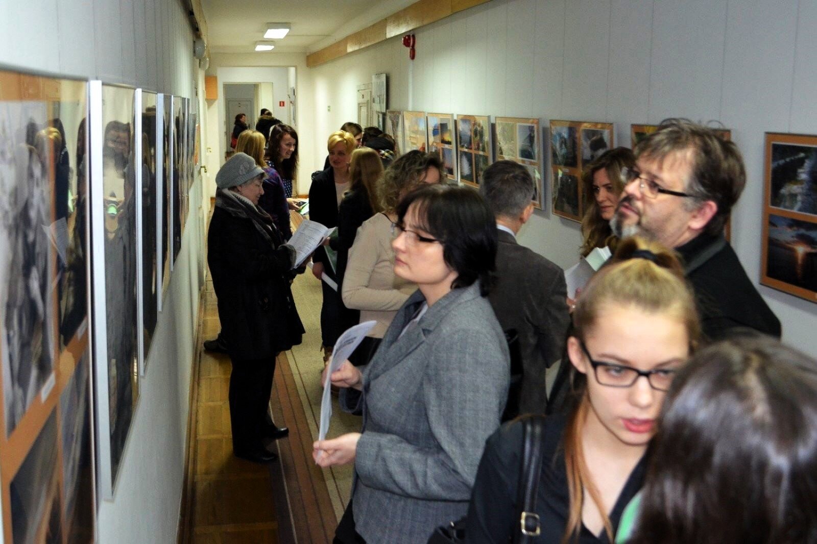 Zdjęcie ukazuje wąski korytarz, na ścianach którego zostały zawieszone fotografie w metalowych ramkach. Wystawę oglądają z dużym zainteresowaniem osoby dorosłe zaproszone na wystawę.
