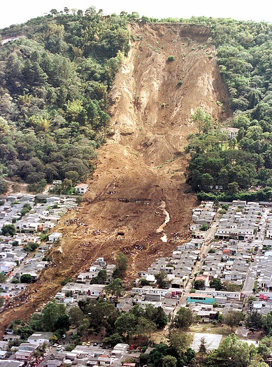 Grafika przedstawia lawinę błotną w Salwadorze, która zeszła na skutek trzęsienia ziemi. Błoto spływa z porośniętego drzewami wzniesienia na liczne zabudowania, które stoją u podnóża wzniesienia. Niektóre zabudowania całkowicie przykryte błotem.  