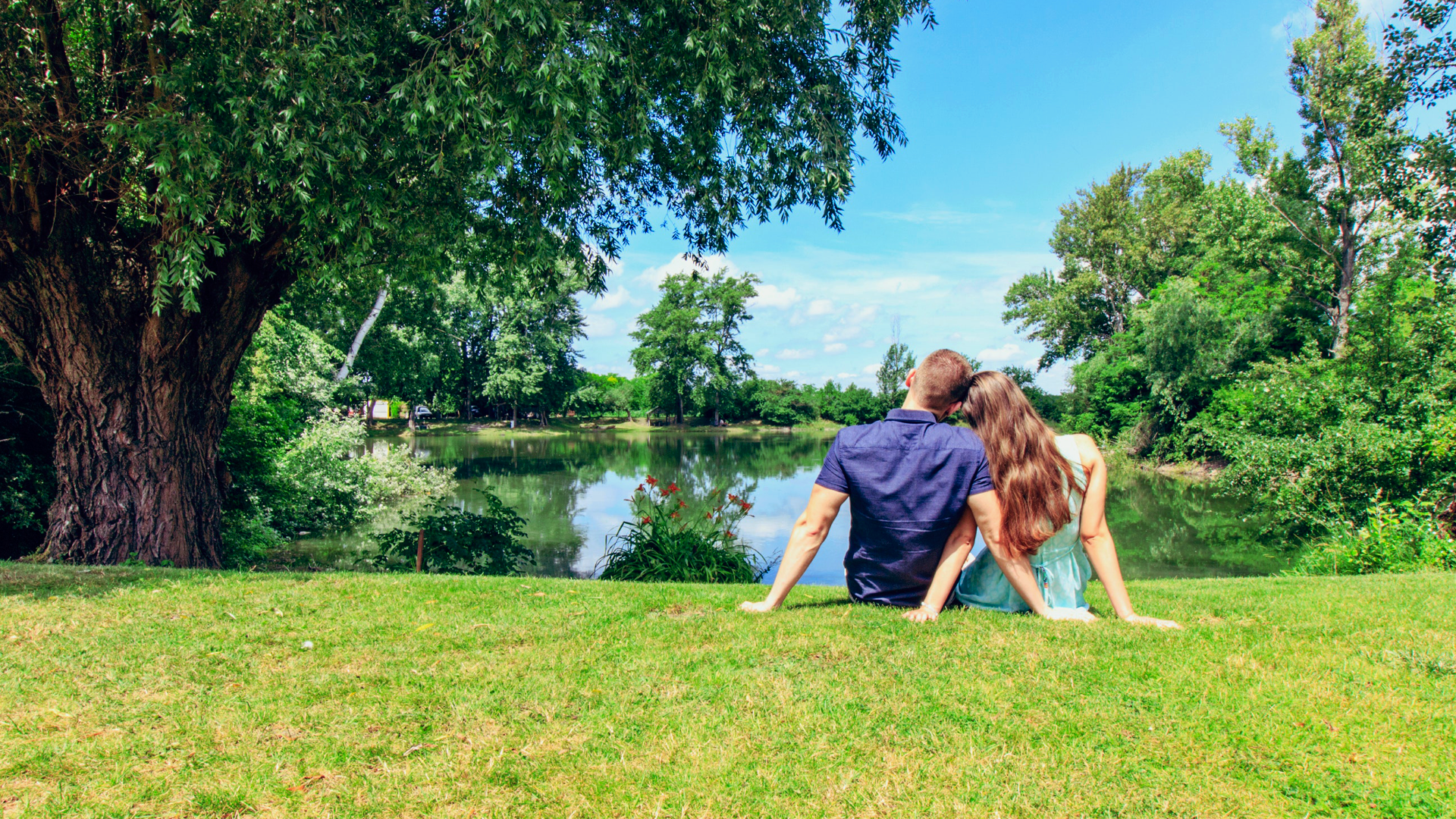 Zdjęcie przedstawia parę, chłopaka i dziewczynę, którzy siedzą na trawie, nad stawem. Chłopak i dziewczyna siedzą tyłem do kadru, bokiem do siebie, są przytuleni do siebie. Patrzą w kierunku stawu. Wokoło jeziora znajdują się drzewa.