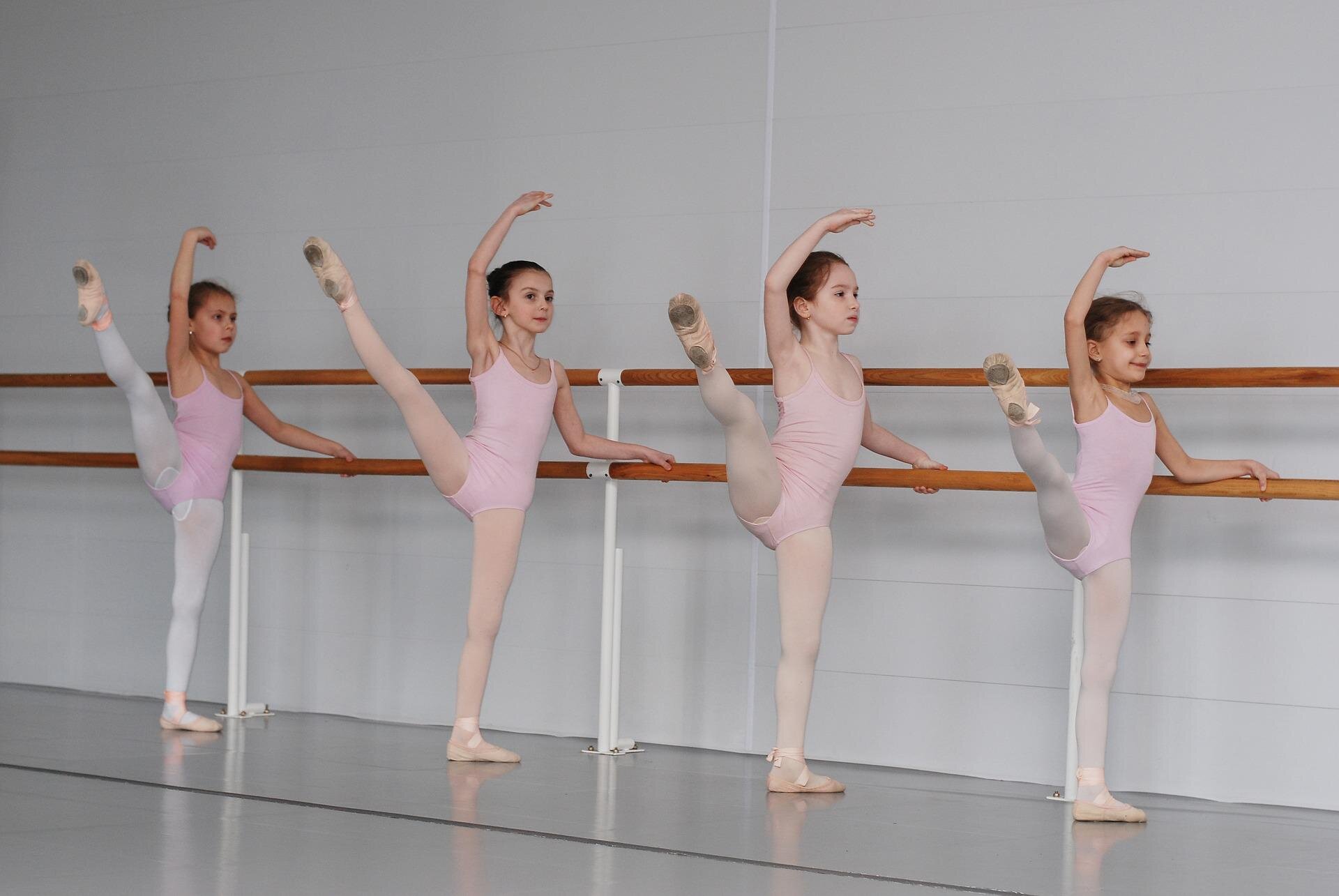 Fotografia przedstawia cztery dziewczynki w rzędzie trzymające się jedną ręką barierki i unoszące jedną nogę na wysokość głowy. Wszystkie ubrane są w różowe stroje do tańca baletu. Na nogach mają założone buty do baletu. 