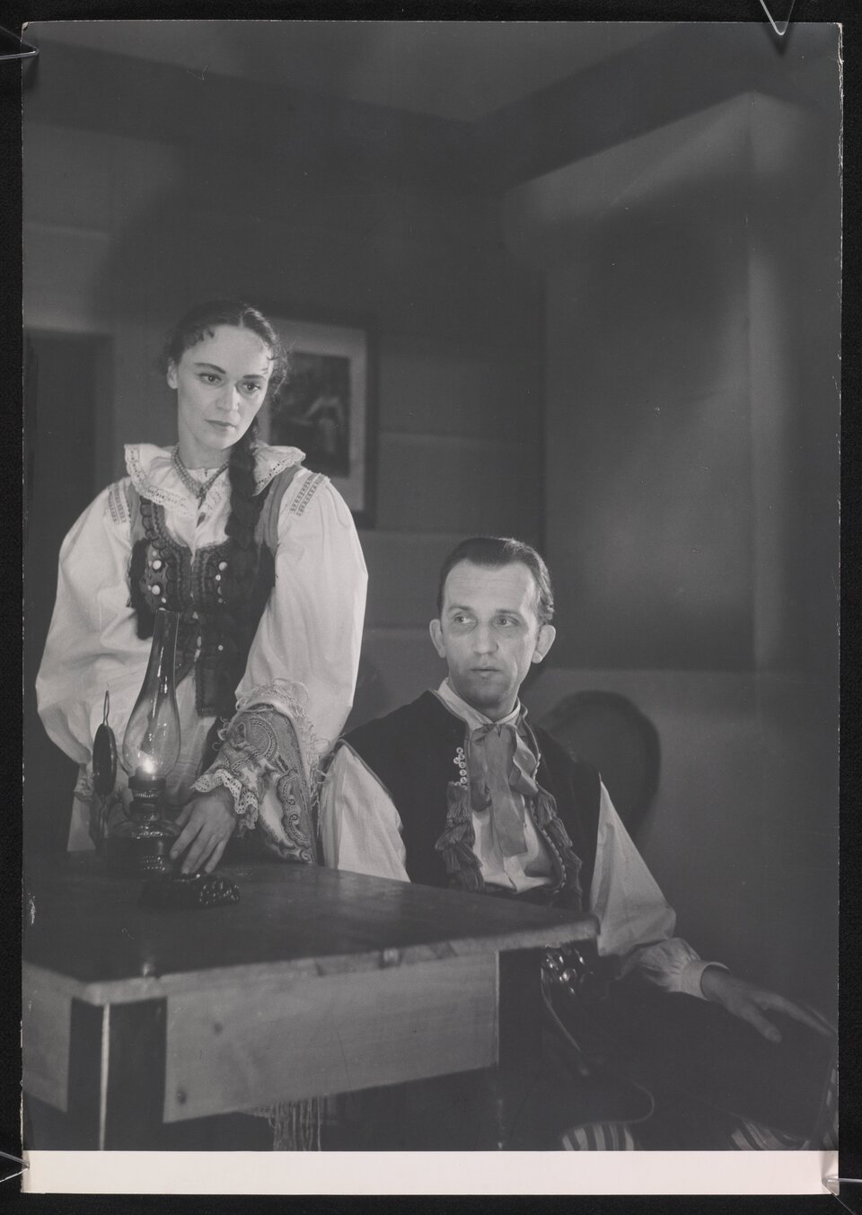 Czarno‑białe zdjęcie przedstawia parę, kobietę i mężczyznę w strojach ludowych. Kobieta stoi przy stole, trzyma znajdującą się na nim lampę. Kobieta patrzy gdzieś przed siebie, ma włosy zaplecione w warkocz, ubrana jest w jasną suknię ze wzorzystym gorsetem. Mężczyzna siedzi na krześle. Patrzy w lewą stronę zdjęcia. Ma krótkie włosy zakolami. Ubrany jest w jasną koszulę, wzorzystą kamizelkę i zawiązaną u szyi chustę. W tle jest ściana z wiszącym obrazem.
