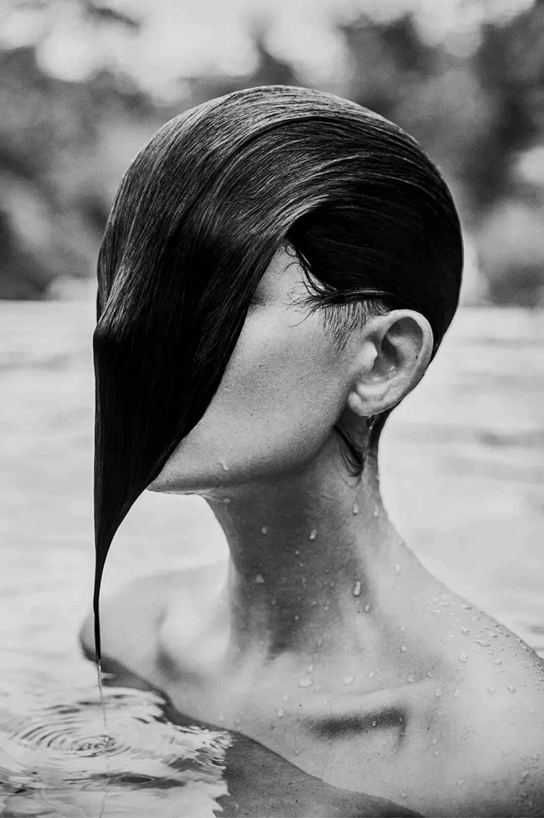 Fotografia przedstawia głowę i nagie ramiona kobiety wynurzającej się z wody. Jej włosy gładko przylegają do głowy, zakrywają całą twarz, ociekają wodą. Kapiący z włosów strumień tworzy kręgi na powierzchni wody.