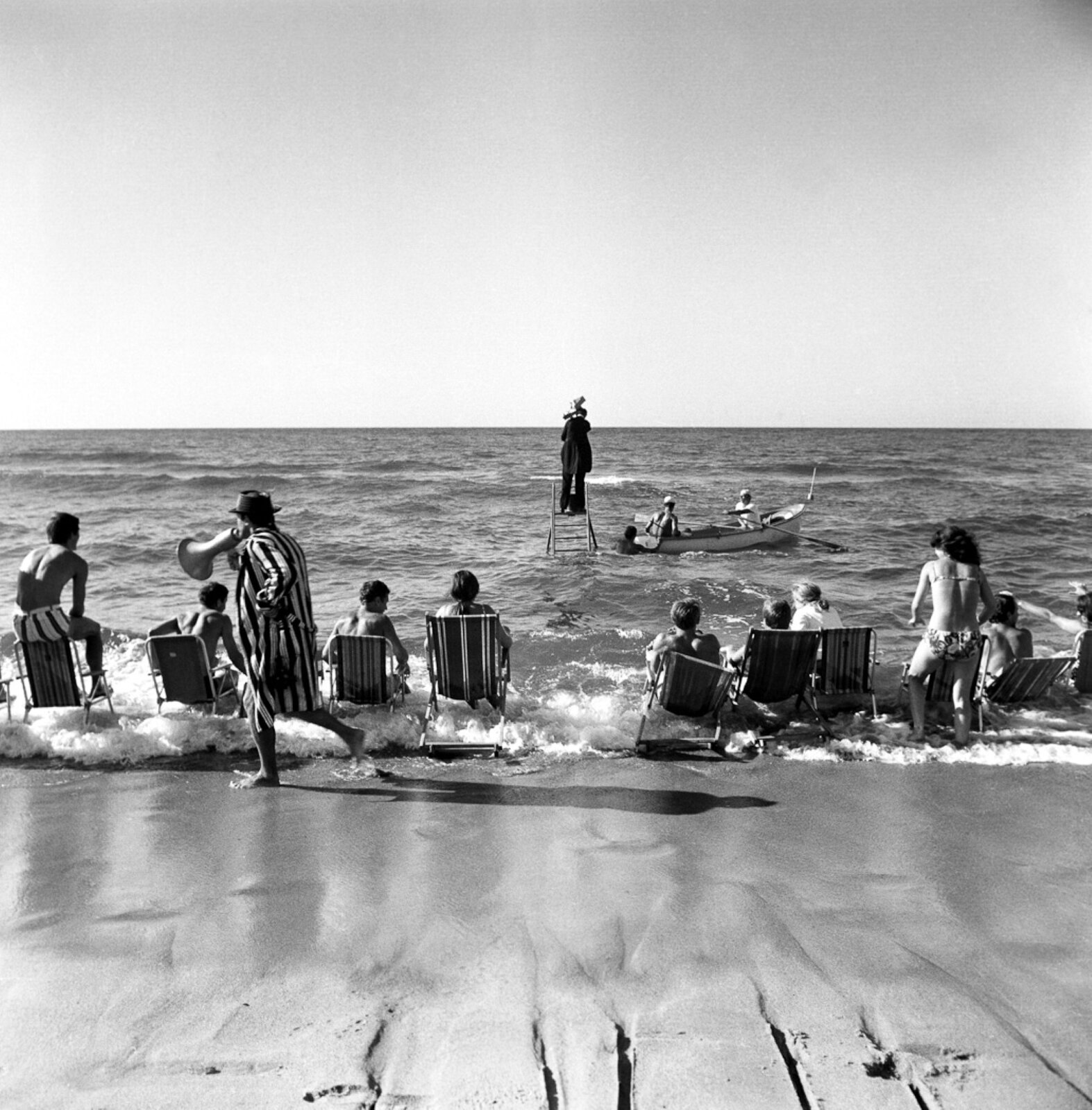 Na starym czarno białym zdjęciu ukazana jest scena rozgrywająca się nad brzegiem morza. W płytkiej wodzie postawiono drabinę, na której stoi ubrany na czarno mężczyzna. Z prawej strony drabiny dryfuje łódka, w której siedzą dwaj mężczyźni trzymający w dłoniach wiosła. Trzeci mężczyzna stoi obok rufy łodzi. Na brzegu ustawiono osiem leżaków, w których siedzą ludzie. Za leżakami przechadza się mężczyzna ubrany w szlafrok w pionowe paski; w dłoni trzyma megafon. 