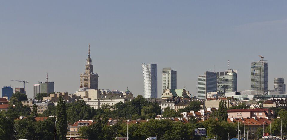Na zdjęciu część śródmieścia Warszawy ujęta ze znacznego oddalenia. Na pierwszym planie zielona roślinność i przebijające spomiędzy niej budynki z ciemnopomarańczowymi dachami. Na drugim planie nowoczesna zabudowa biurowa. Bardzo wysokie budynki w centrum miasta – głównie wieżowce, a po lewej stronie kadru wieża Pałacu Kultury i Nauki. W tle szare bezchmurne niebo.