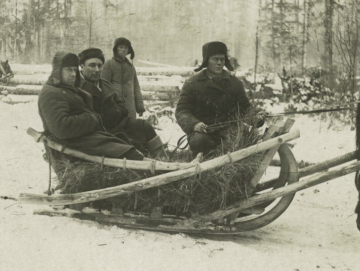 Zdjęcie przedstawia trzech mężczyzn jadących w drewnianych saniach wyściełanych sianem. Jeden z nich trzyma w rękach lejce. Za saniami stoi czwarty mężczyzna. Wszyscy ubrani są w grube płaszcze i czapki uszatki. Na dalszym planie są ścięte pnie drzew i las.