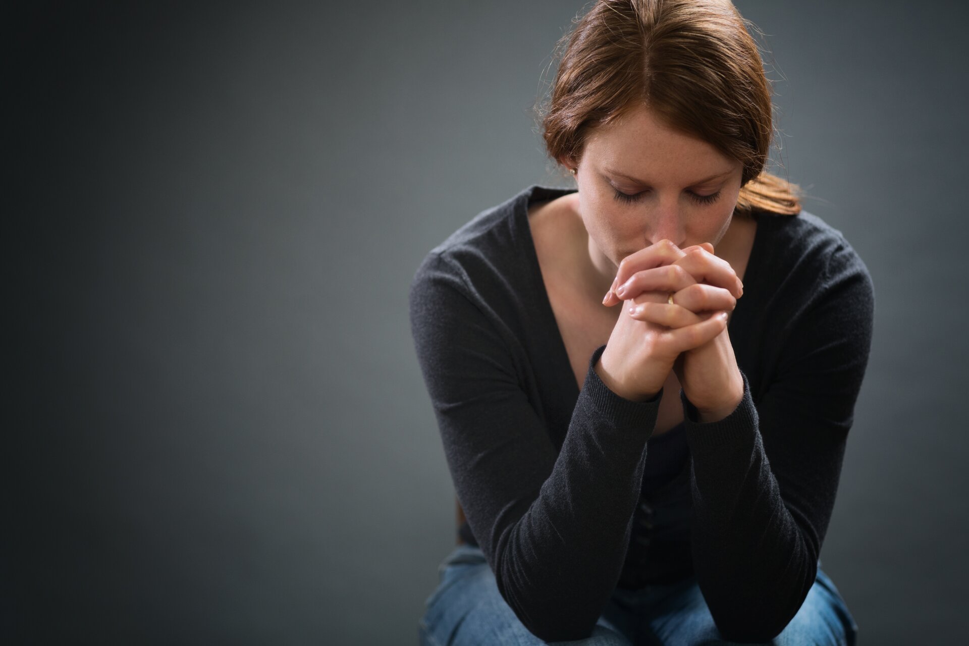 Ilustracja przedstawia kobietę ubraną w ciemną bluzkę oraz sweter. Kobieta ma długie ciemne włosy, związane w kucyk. Postać siedzi lekko pochylona do przodu. Ma zamknięte oczy. Jej łokcie spoczywają na kolanach, a brodę opiera na dłoniach.