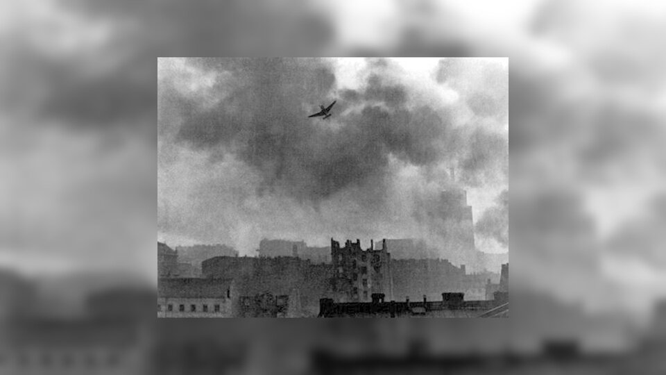 Zdjęcie przedstawia panoramę zbombardowanej części miasta. Nad ruinami budynków unoszą się samolot oraz kłęby dymu.
