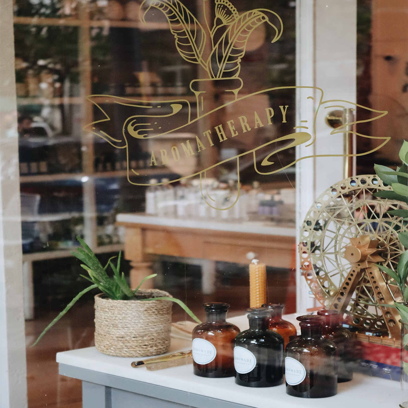Zdjęcie przedstawia witrynę sklepu. Na szybie widoczny jest napis w języku angielskim: AROMATHERAPY. Na wystawie widoczne są butelki z brązowego szkła oraz doniczka z kwiatem. 