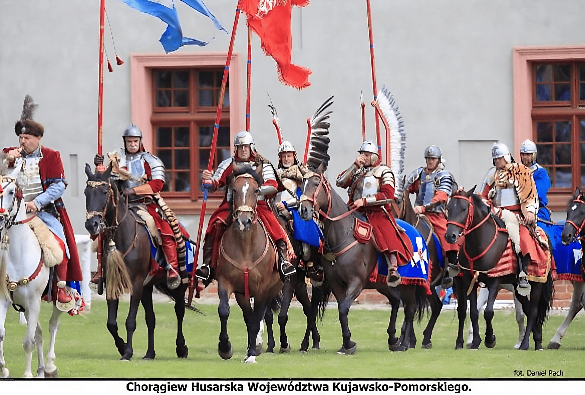 Ilustracja przedstawia grupę ludzi na koniach, ubranych w stroje husarskie, ujętych z przodu. Mężczyźni mają na sobie zbroje, hełmy i czerwone lub niebiesko‑czerwone okrycia wierzchnie. Niektórzy mają założone na plecy husarskie skrzydła, a w rękach trzymają czerwone piki z powiewającymi na ich końcach czerwonymi i niebiesko‑białymi proporcami. Jeden z mężczyzn, widoczny po lewej stronie kadru, jest ubrany w purpurowy płaszcz, spod którego wystaje błękitna szata, a na głowie ma czapkę z broszką w części czołowej, nad którą znajdują się poóra. Konie mają założone niebieskie czapraki z czerwonymi obszyciami oraz białymi krzyżami. W tle widać fragment budynku z białą elewacją, z dużymi oknami o brązowych ramach.