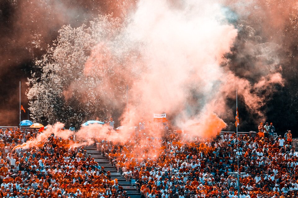 Zdjęcie przedstawia trybunę stadionu wypełnioną kibicami. Nad trybuną unosi się dym z rac.