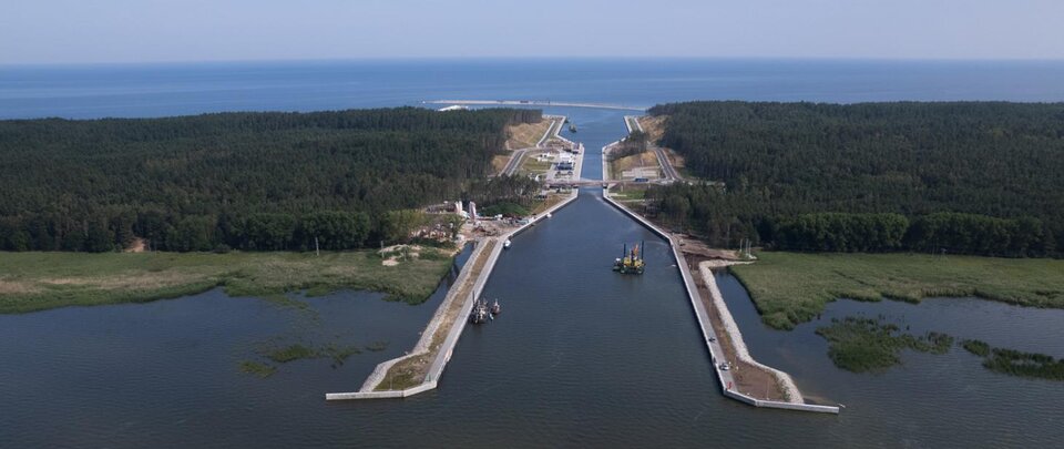 Kanał wodny przekopany przez zalesioną mierzeję prowadzi od zalewu do otwartego morza. Po obu stronach kanału rozciągają się lasy i tereny podmokłe. W tle widoczna jest linia brzegowa oraz spokojna tafla morza.