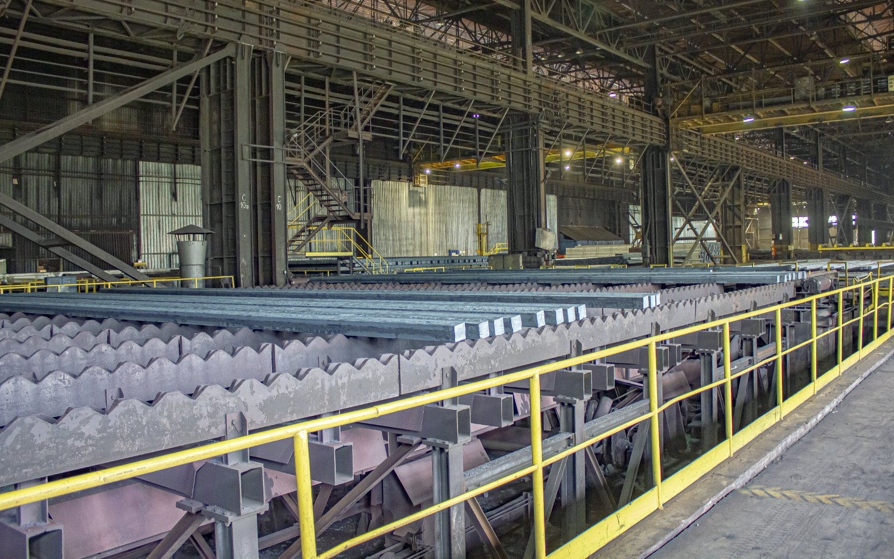 Fotografia przedstawia halę produkcyjną na której chłodzi się odlane wlewki, czyli długie pręty o prostokątnym przekroju. Na obrazku widać paręnaście wlewków na urządzeniu transportowym, składającym się z ząbkowanych blach umieszczonych tak, że przekrój prętów jest równoległy do płaszczyzny blach, a krawędzie wlewków idealnie pasują do krawędzi ząbków blachy. Przesuwając odpowiednio blachy, wlewki przesuwane są dalej po linii produkcyjnej.