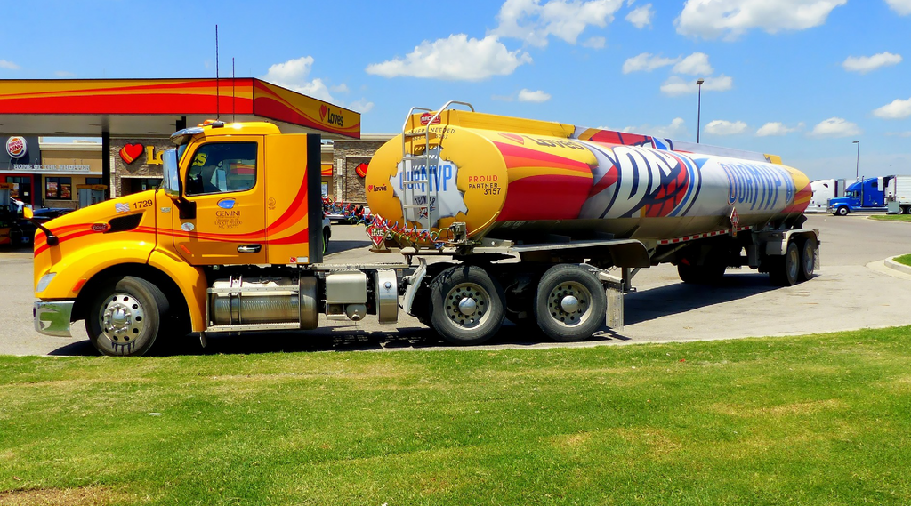 Na zdjęciu widoczny jest pojazd cysterna o podwoziu cylindrycznym. Cysterna na podwoziu samochodowym jest w kolorze żółto‑czerwonym. Na beczce cysterny znajduje się grafika ozdobna. Beczka cysterny jest o przekroju elipsy. W tle zdjęcia widać stacje paliwową.