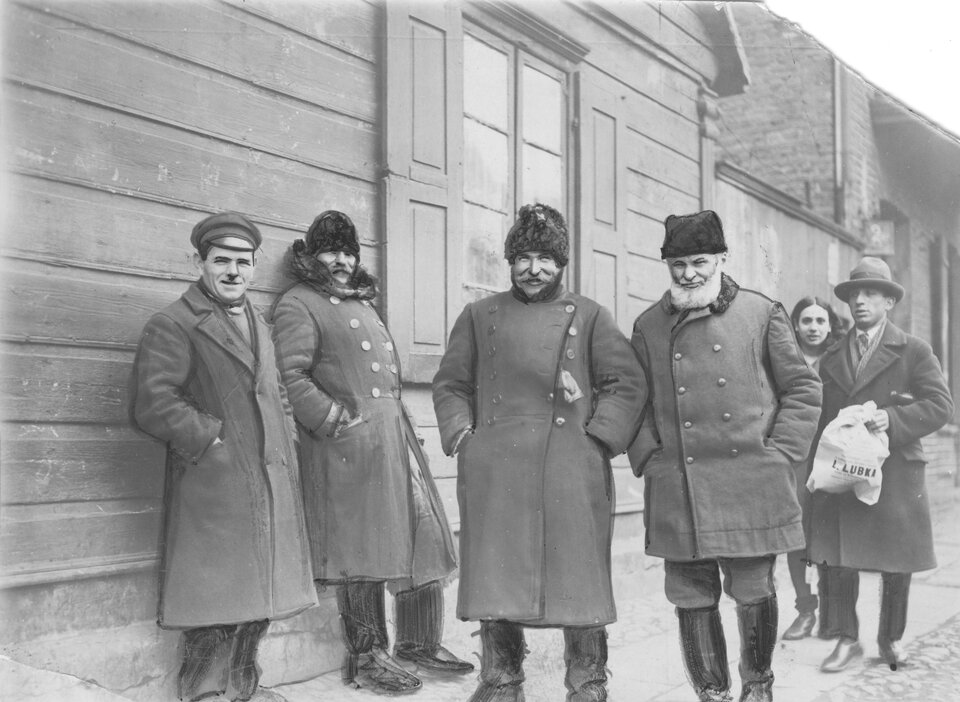 Ilustracja przedstawia grupę mężczyzn. Jest ich sześciu. Wszyscy mają na sobie szare długie płaszcze. Pięciu ma na głowach czapki. Jeden z nich ma gęstą białą brodę. Inny trzyma w ręce reklamówkę. Stoją na tle drewnianej chaty, pod okiennicą.