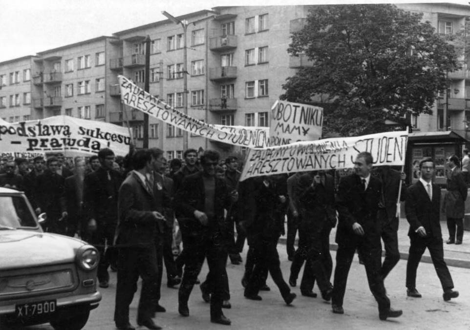 Czarno-biała fotografia przedstawiająca wrocławskich studentów w czasie pochodu pierwszomajowego w 1968 roku, którzy domagali się uwolnienia aresztowanych kolegów. Demonstranci niosą transparenty z napisami:  Podstawa szacunku – prawda; Żądamy uwolnienia aresztowanych studentów.
