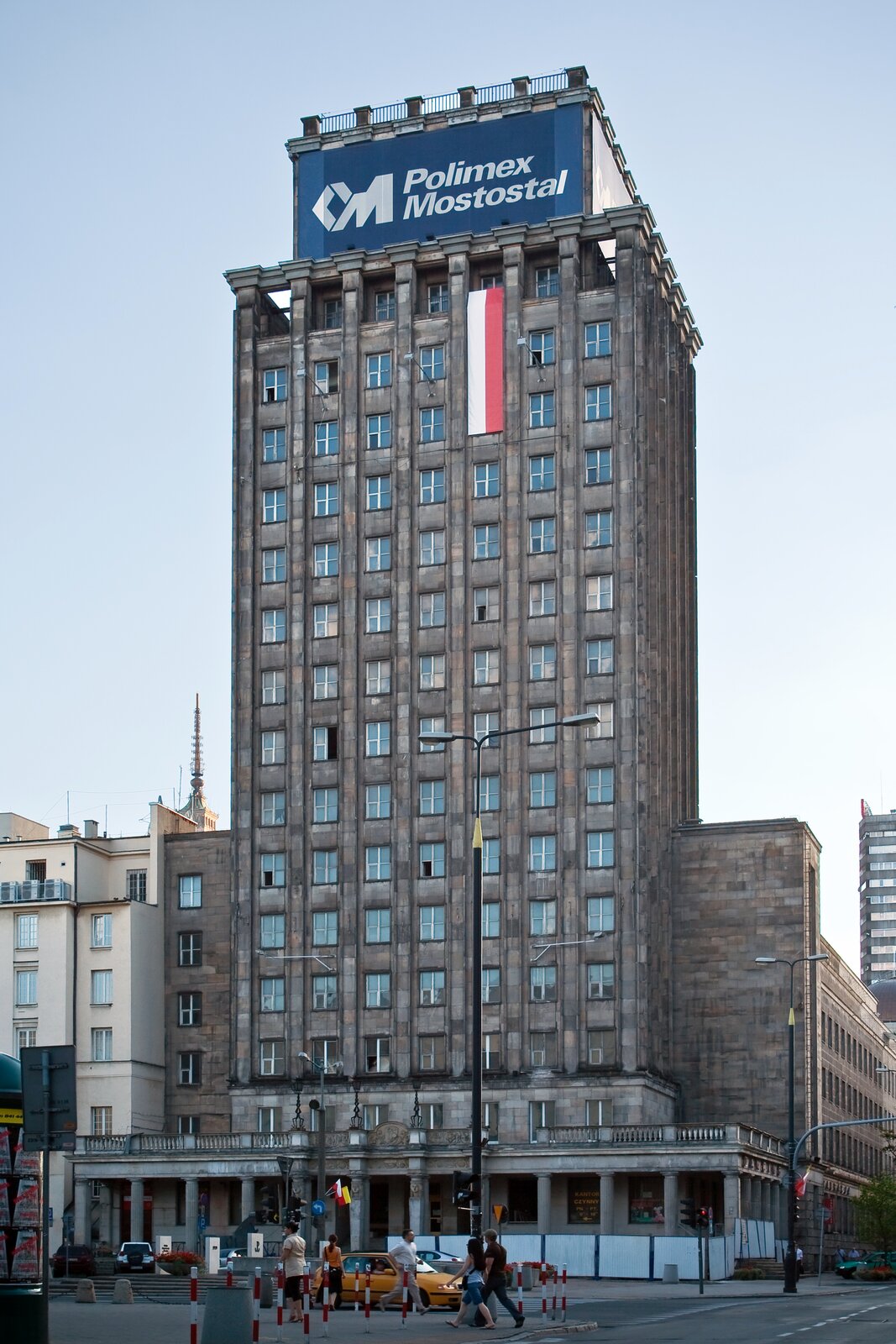 Rys. 14. Zdjęcie prezentuje budynek Prudential (obecnie Hotel Warszawa) na placu Napoleona (obecnie Plac Powstańców Warszawy), który mieścił pierwsze w Polsce studio telewizyjne. Zdjęcie pochodzi z dwa tysiące ósmego roku. Kilkunastopiętrowy ciemnoszary budynek zdobi pionowo zawieszona długa na kilka pięter flaga Polski. Na szczycie budynku rozpościera się reklama. Na granatowym tle widać napis i logo firmy Polimex Mostostal. 