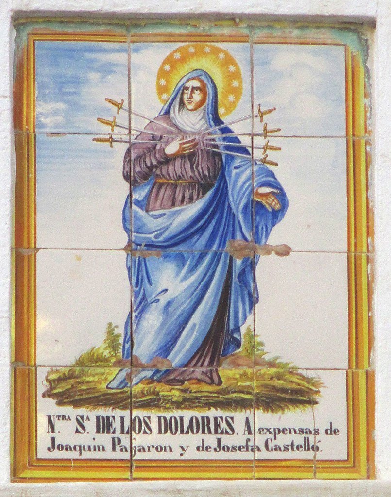 Zdjęcie przedstawia stworzony z kafli ołtarz, który przedstawia stojącą postać Matki Boskiej. To dojrzała kobieta w długiej sukni, okryta na głowie i ramionach zwojem materiału. Nad jej głową znajduje się rozświetlona promieniami i gwiazdami aureola. Kobieta ma wbitych siedem długich sztyletów w klatkę piersiową. Trzyma na piersiach prawą dłoń. Pod obrazem znajduje się napis w języku hiszpańskim.