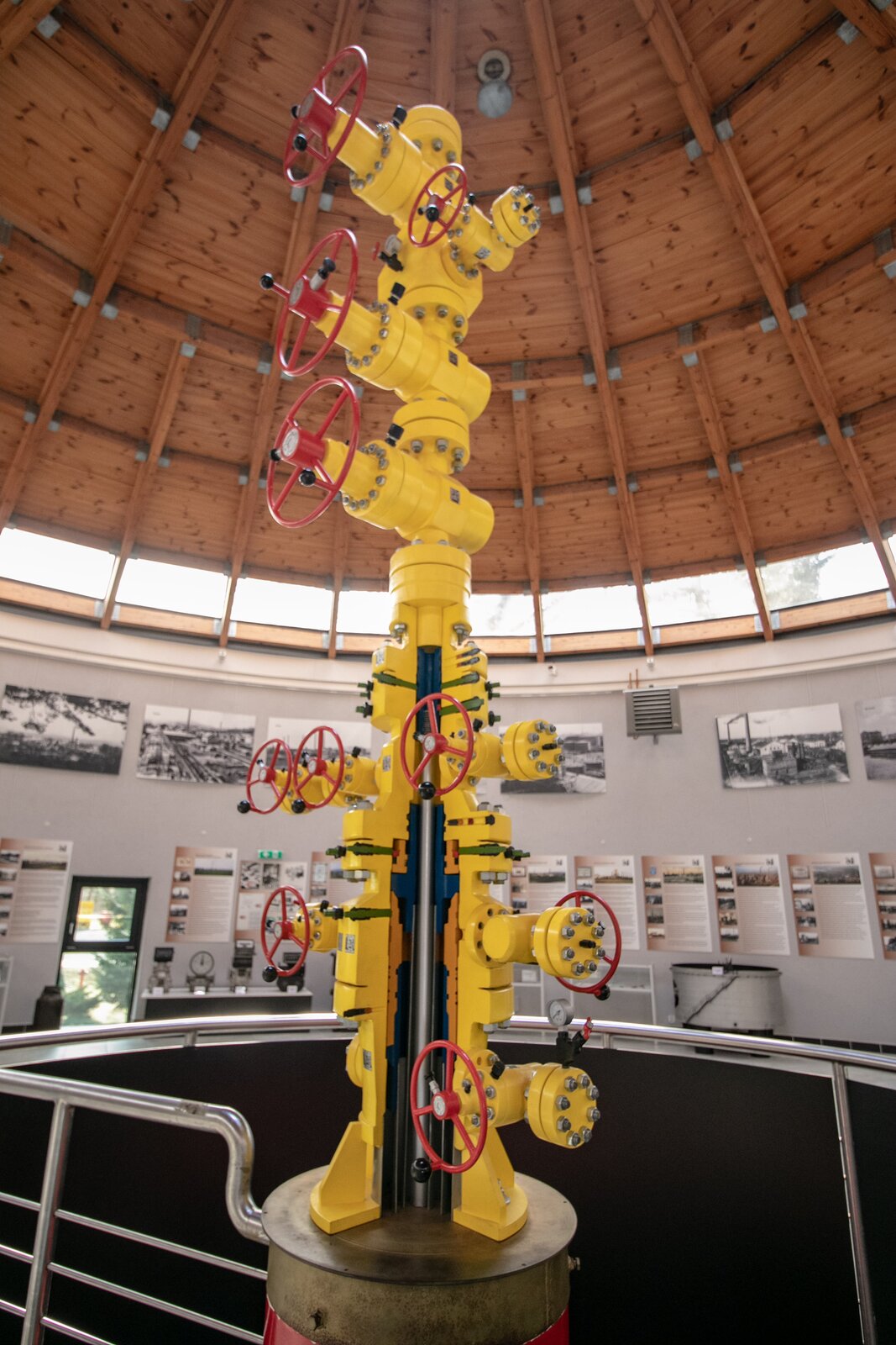 Na pionowym zdjęciu widoczna jest głowica eksploatacyjna umiejscowiona w okrągłym pomieszczeniu z drewnianym dachem oraz widocznymi na ścianie za głowicą zdjęciami i grafikami, które są nieczytelne. Głowica eksploatacyjna jest wydłużonym elementem o żółtym korpusie, składającym się z połączonych ze sobą zasów posiadających koła koloru czerwonego do ich obsługi. W górnej części głowicy eksploatacyjnej widoczne są 3 koła zwrócone ku przodowi oraz 1 mniejsze koło, umiejscowione z boku poniżej pierwszego zaworu od góry. W dolnym fragmencie głowicy widoczny jest półprzekrój głowicy jako wycinek 1/4 części głowicy na połowie jej długości od dołu. W przekroju widoczne są liczne zwężenia przekroju oraz srebrny wydłużony element zlokalizowany pośrodku. W górnej części przekroju widoczne trzy zawory zlokalizowane poziomo, przy czym dwa koła zlokalizowane są po stronie lewej, a jedno po prawej. Poniżej widoczne dwa koła symetrycznie usytuowane, jednak lewe skierowane jest ku przodowi, a prawe ku tyłowi modelu. Poniżej nich widoczne większe koło zlokalizowane z prawej strony. Cały model znajduje się na okrągłym postumencie, od którego w lewą stronę odchodzi balustrada o okrągłym kształcie. Głowica eksploatacyjna ma za zadanie szczelne zamknięcie odwiertu, które zapewnia całkowite zamknięcie lub całkowite otwarcie odwiertu. 