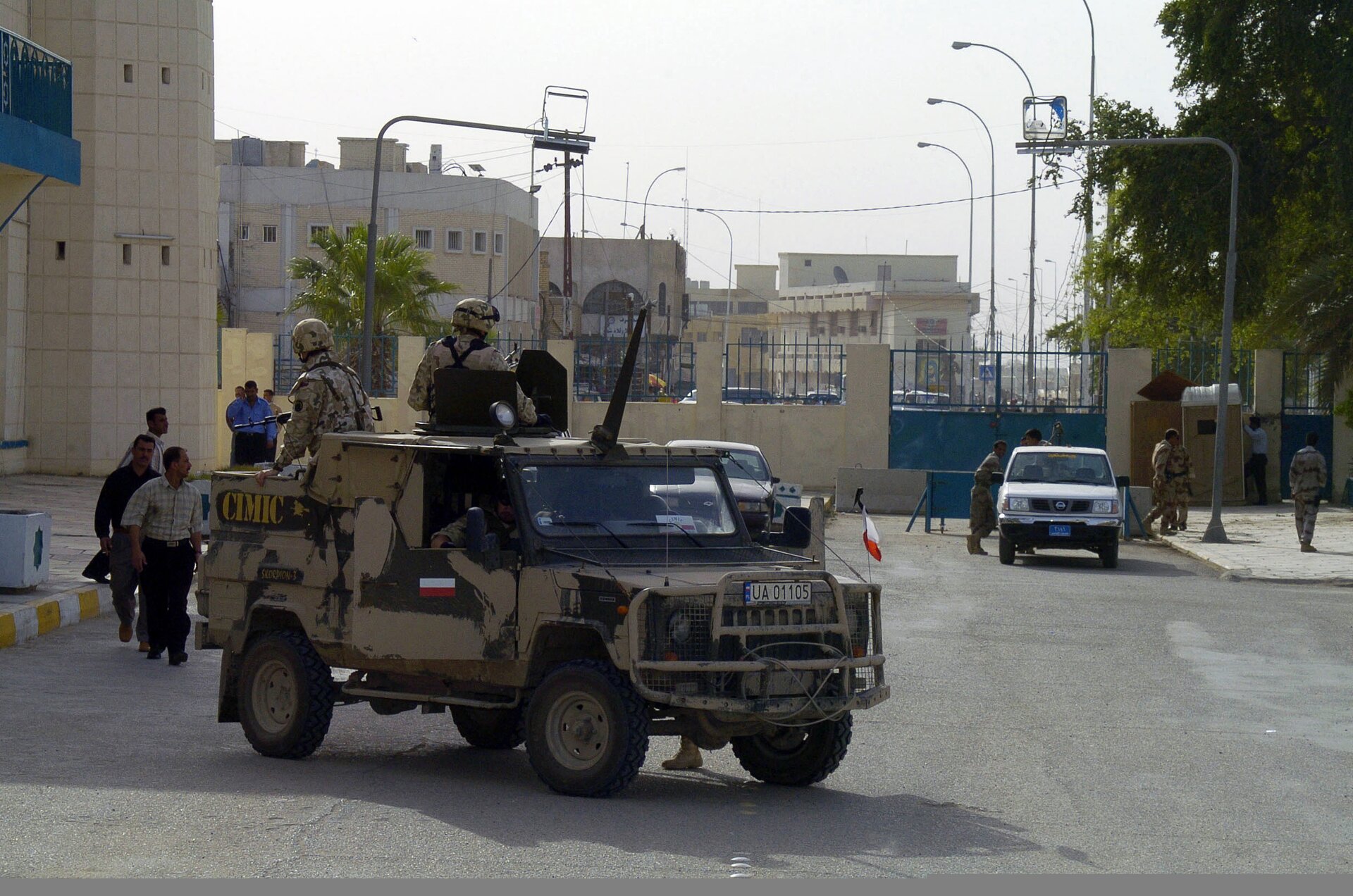 Na zdjęciu znajduje się polski patrol w samochodzie wojskowym w Iraku. Samochód na zdjęciu znajduje się bliżej lewej krawędzi fotografii. Jest opancerzony i posiada dwa otwory - jeden z tyłu samochodu dla strzelca z karabinem, drugi nad kabiną pojazdu dla obserwatora/drugiego strzelca. Dwóch żołnierzy z obu punktów wozu obserwuje okolicę, będąc odwróconymi do widza. Żołnierze i pojazd są maskowane w kolorze pustynnym. W tle utrwalona jest zabudowa miasta.