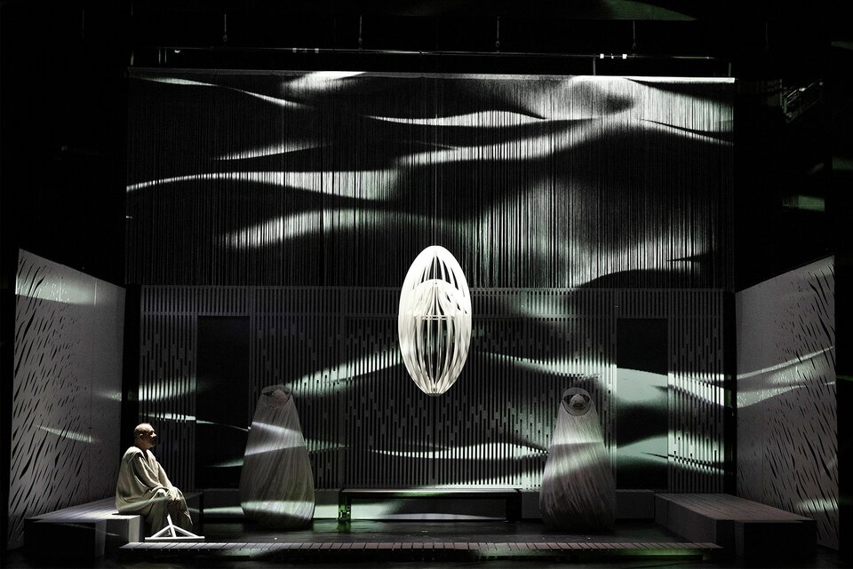 Fotografia przedstawia scenografię do przedstawienia teatralnego. Scenografia składa się z białych ścianek, przy których ustawione zostały dwie figury owinięte w jasny materiał i białe ławki. Na jednej z nich siedzi starszy mężczyzna ubrany w białą szatę. Na środku pomieszczenia wisi duży biały żyrandol.