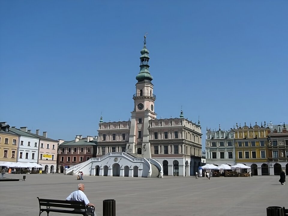 Zdjęcie przedstawia rynek w Zamościu. W centrum widoczna konstrukcja renesansowego ratusza, z wysokim przedprożem i schodami prowadzącymi do wysokiego parteru. Pod schodami pełnołukowy portal. Fasada budynku utrzymana w stylu włoskim - równe odstępy między prostokątnymi oknami, porządkowanie fasady za pomocą fryzów i gzymsów. W centrum szerokie pilastry zwężają się ku górze, aż do wieży zegarowej ratusza z hełmem. Ratusz jest czterokondygnacyjny. Po prawej stronie fotografii Kamienice Ormiańskie czterokondygnacyjne, w parterze mają arkady. Na górze bogato zdobiona attyki. Jeden budynek jest biały, drugi żółty. Po lewej stronie kamienice w pierzei rynku. Przed kamienicami stoją kawiarniane parasole. W tle bezchmurne niebo.