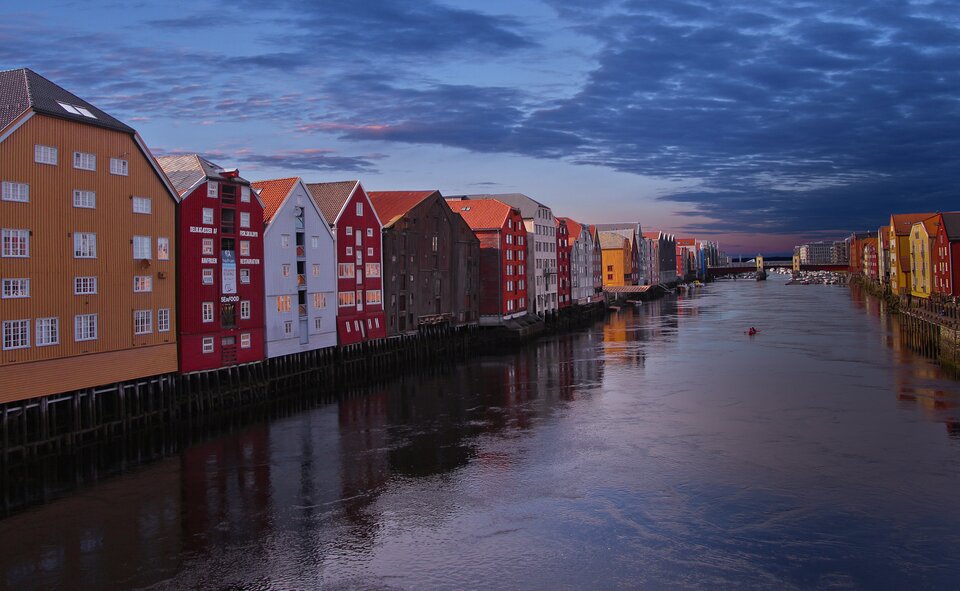 Zdjęcie ukazuje kolorowe, kilkukondygnacyjne domy. Domy mają spadziste dachy, kolorowe fasady, między innymi białe, czerwone, pomarańczowe. Budynki stoją na palach po dwóch stronach rzeki. W tle znajduje się most przez rzekę.    
