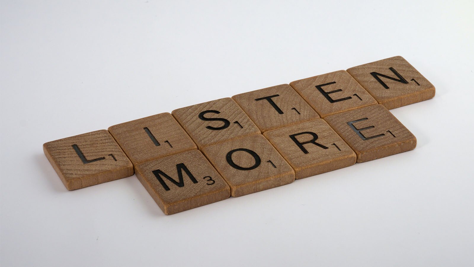 Zdjęcie przedstawia drewniane klocki z literkami do gry, ułożone w dwóch rzędach na białym tle. U dołu, po prawej stronie każdej literki widoczna jest mała cyfra. Klocki układają się w napis w języku angielskim: LISTEN MORE.